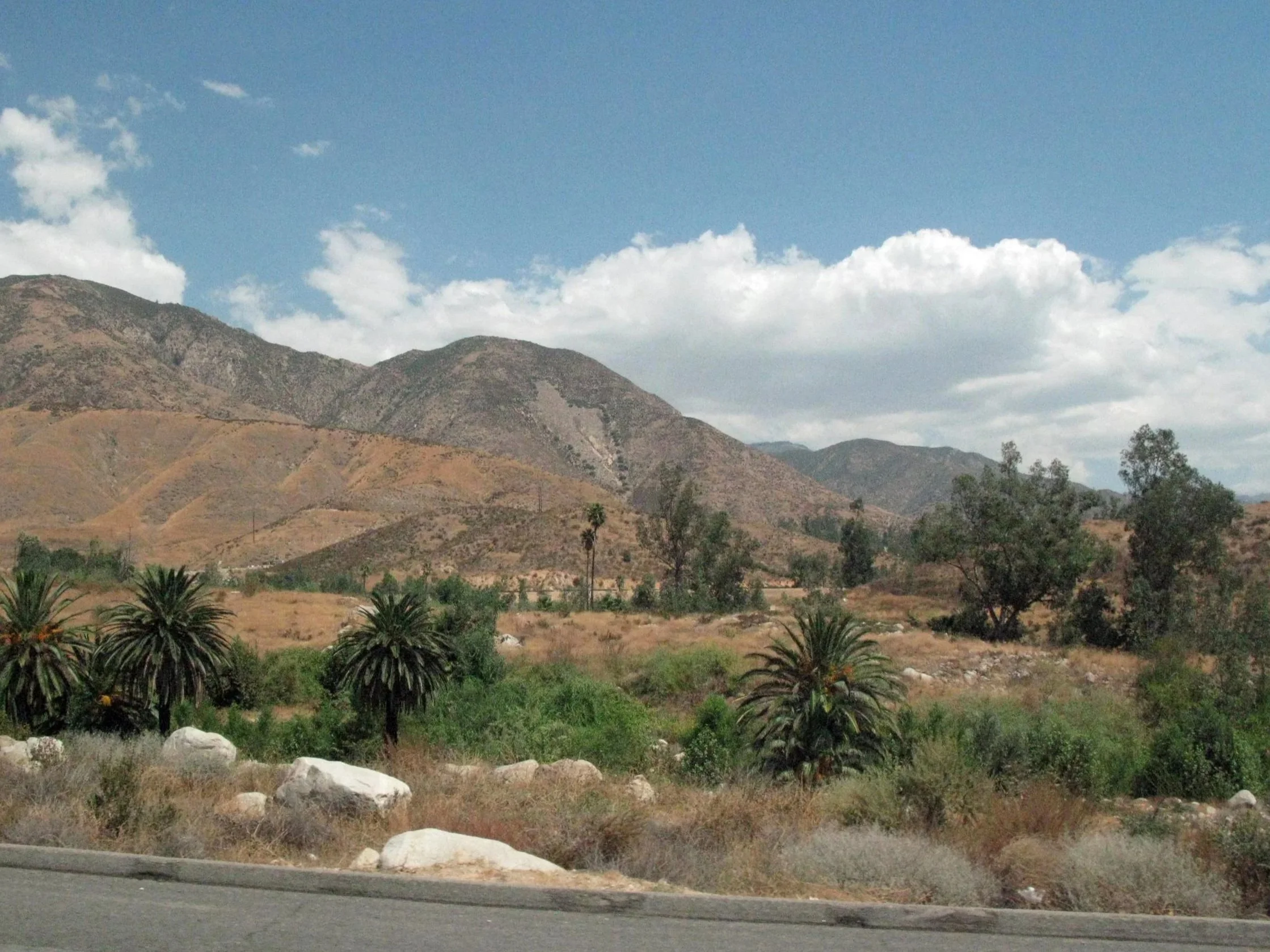 Arrowhead in the San Bernardino Mountains. Hilly landscape with mountains, trees, bushes, and a partly cloudy sky.