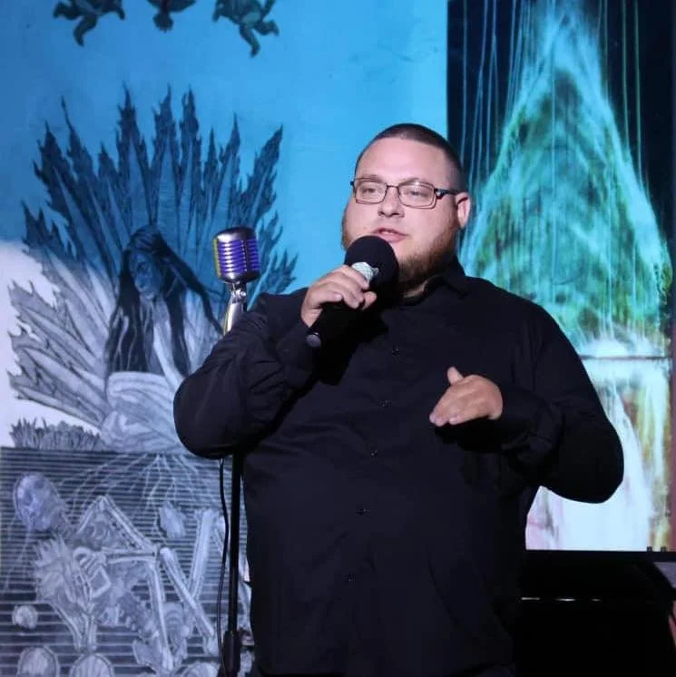 Kyle Carignan. A man with glasses and a beard wearing a black shirt, holding a microphone and performing on stage with a colorful abstract background.