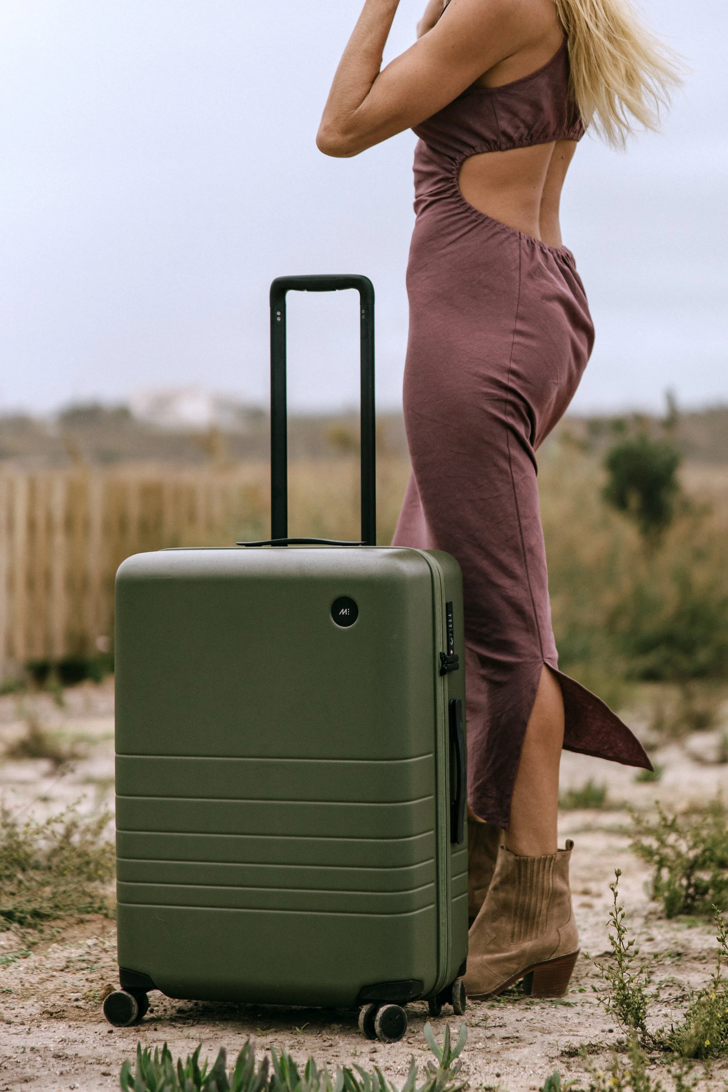 A woman standing outdoors with a green suitcase on wheels, wearing a sleeveless dress and ankle boots in Ericeria, Portugal