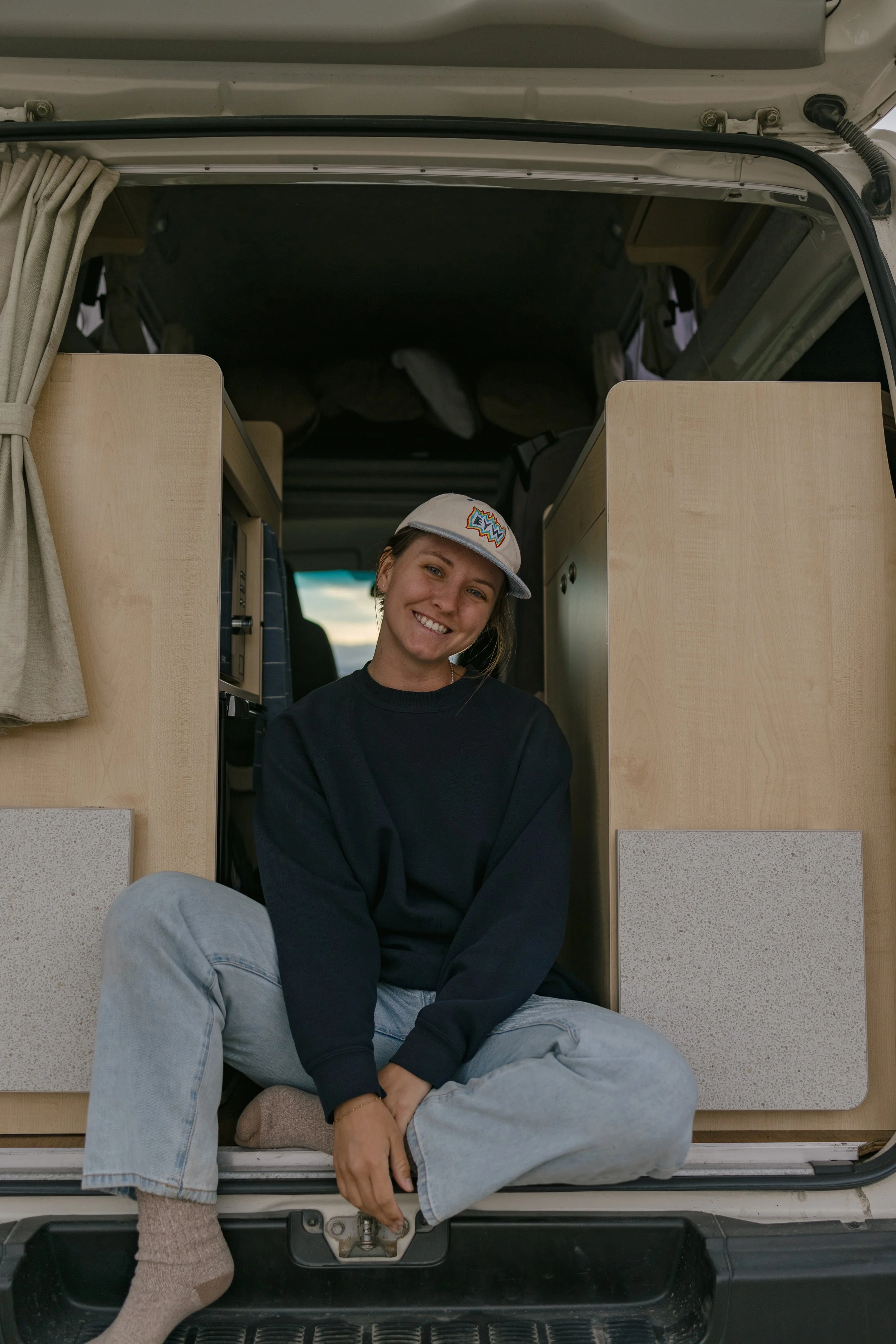 A young woman sitting in the open back of a camper van, smiling at the camera. She is wearing a beige baseball cap, a black sweatshirt, and light jeans in New Zealand