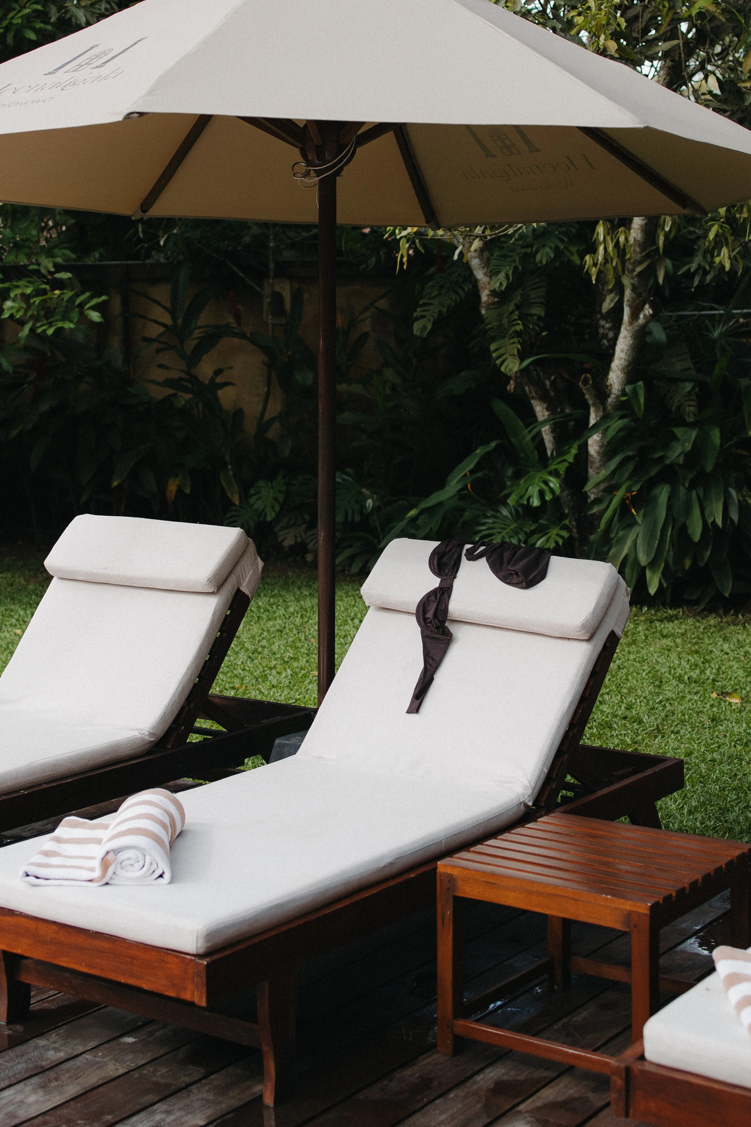 Two white outdoor lounge chairs with rolled towels on a wooden deck, shaded by a large beige patio umbrella, surrounded by green plants and trees in Sri Lanka