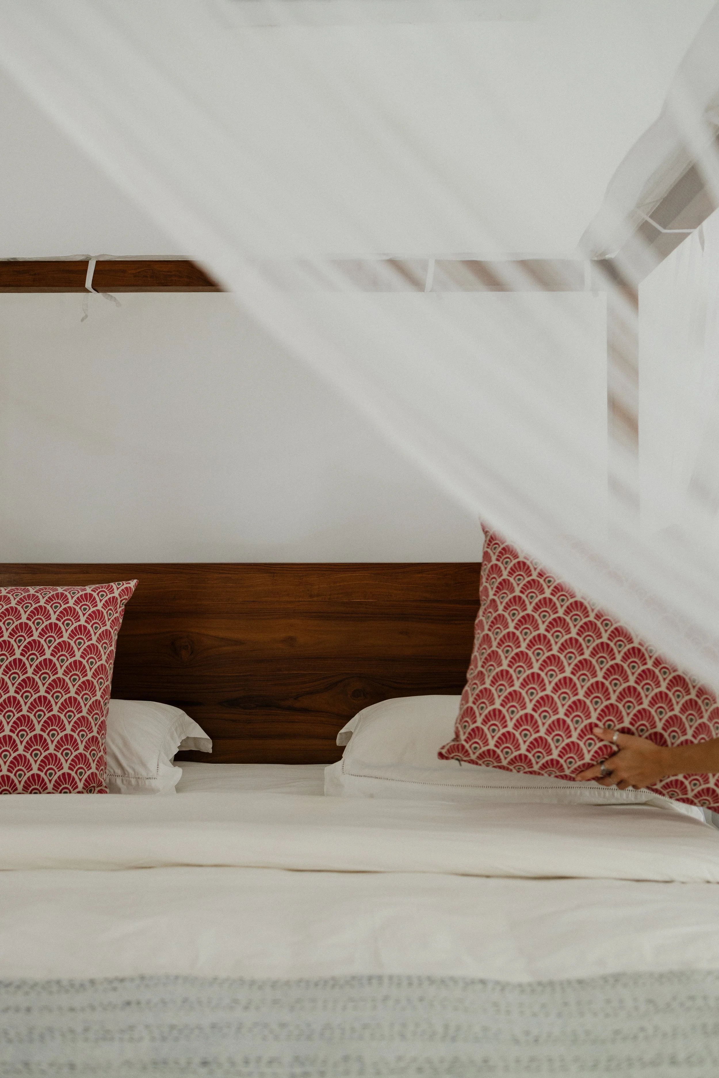 A neatly made bed with white bedding and two pillows with red and white patterned pillowcases, against a wooden headboard. A person's hand is visible placing a similar patterned pillow onto the bed in an Airbnb in Sri Lanka