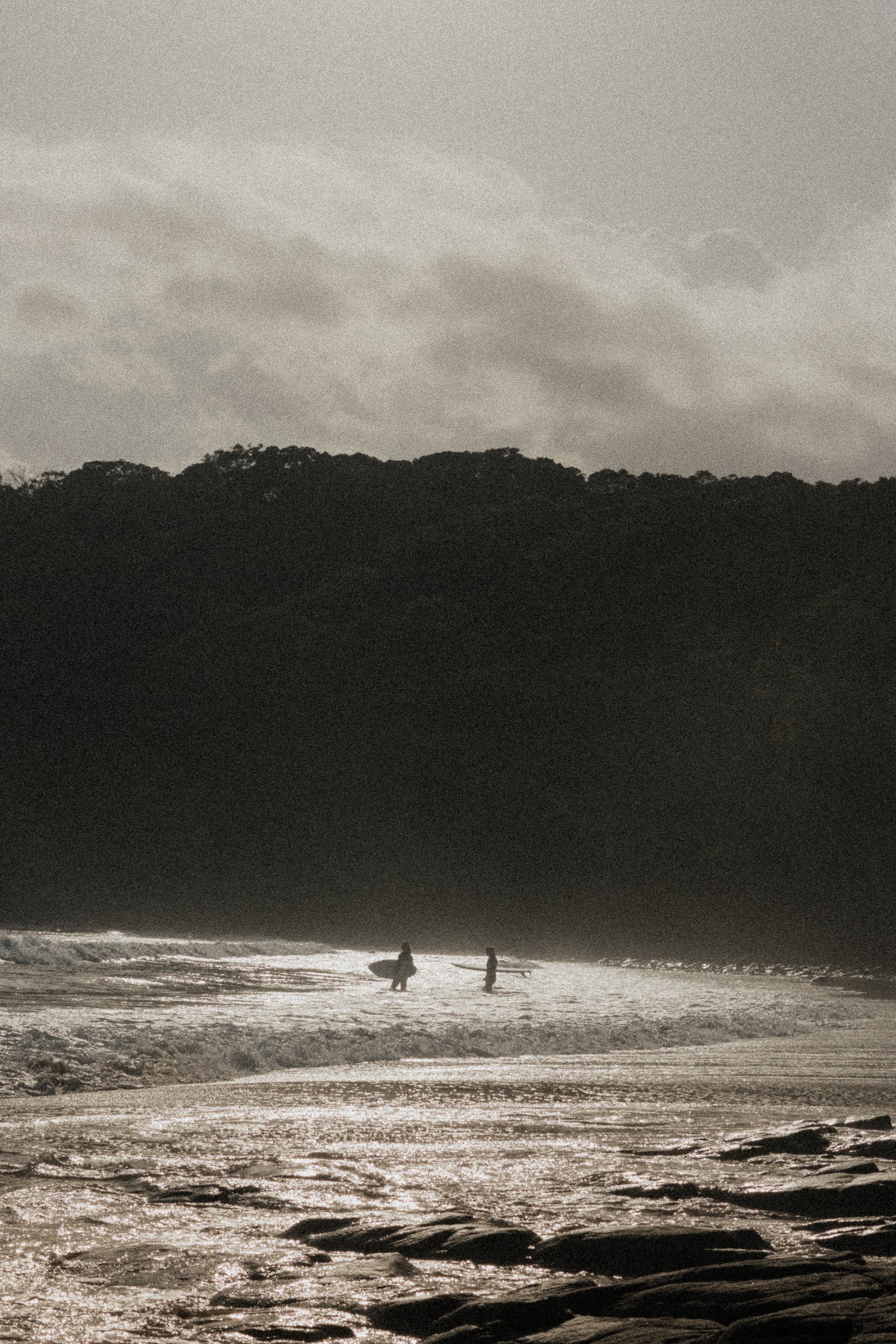 Two surfers walking along the beach near the water, with a forested hillside in the background and a cloudy sky overhead in Sunshine Coast, Australia