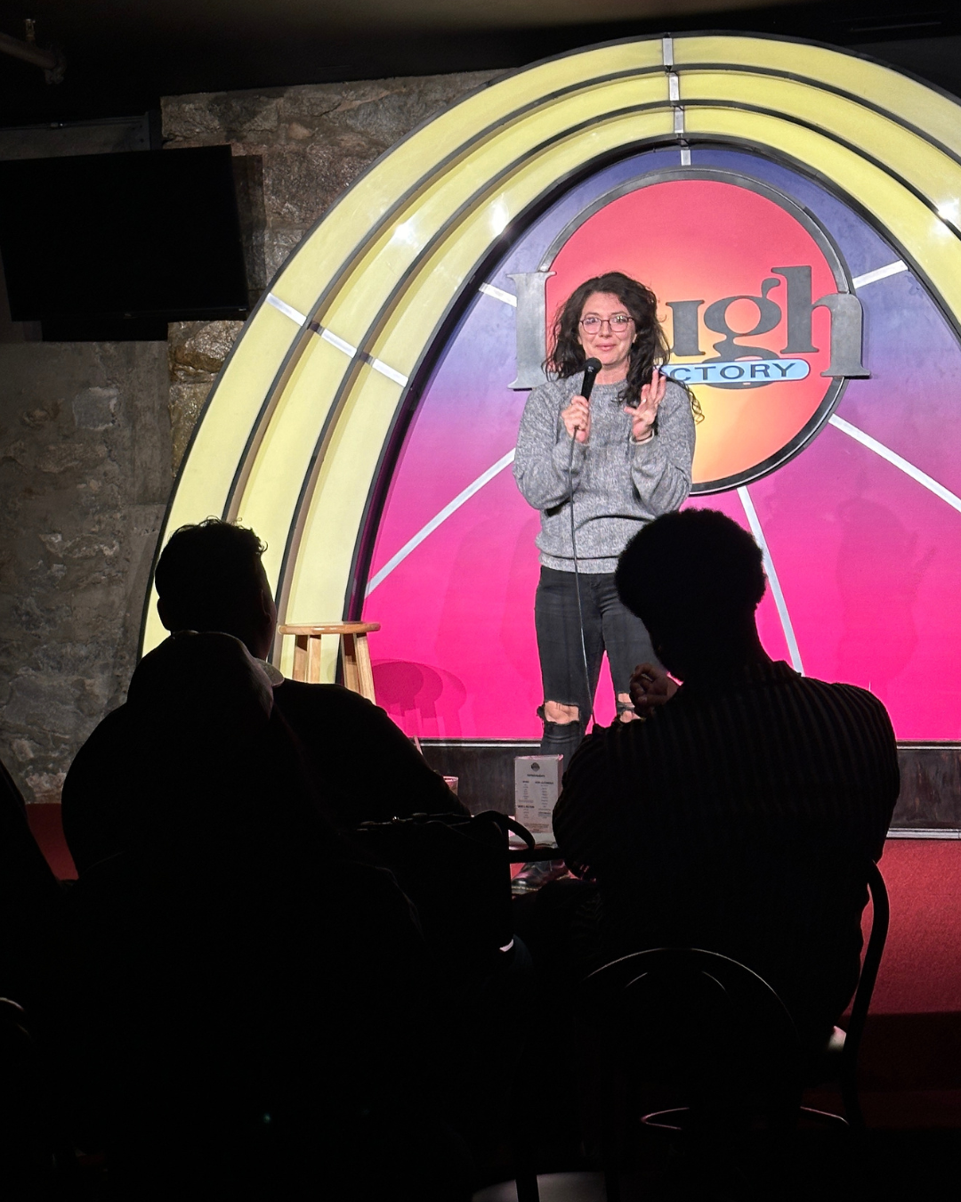 A woman stand-up comedian performing on stage at the Laugh Factory, holding a microphone, with a colorful Laugh Factory backdrop behind her, and audience members in silhouette watching.