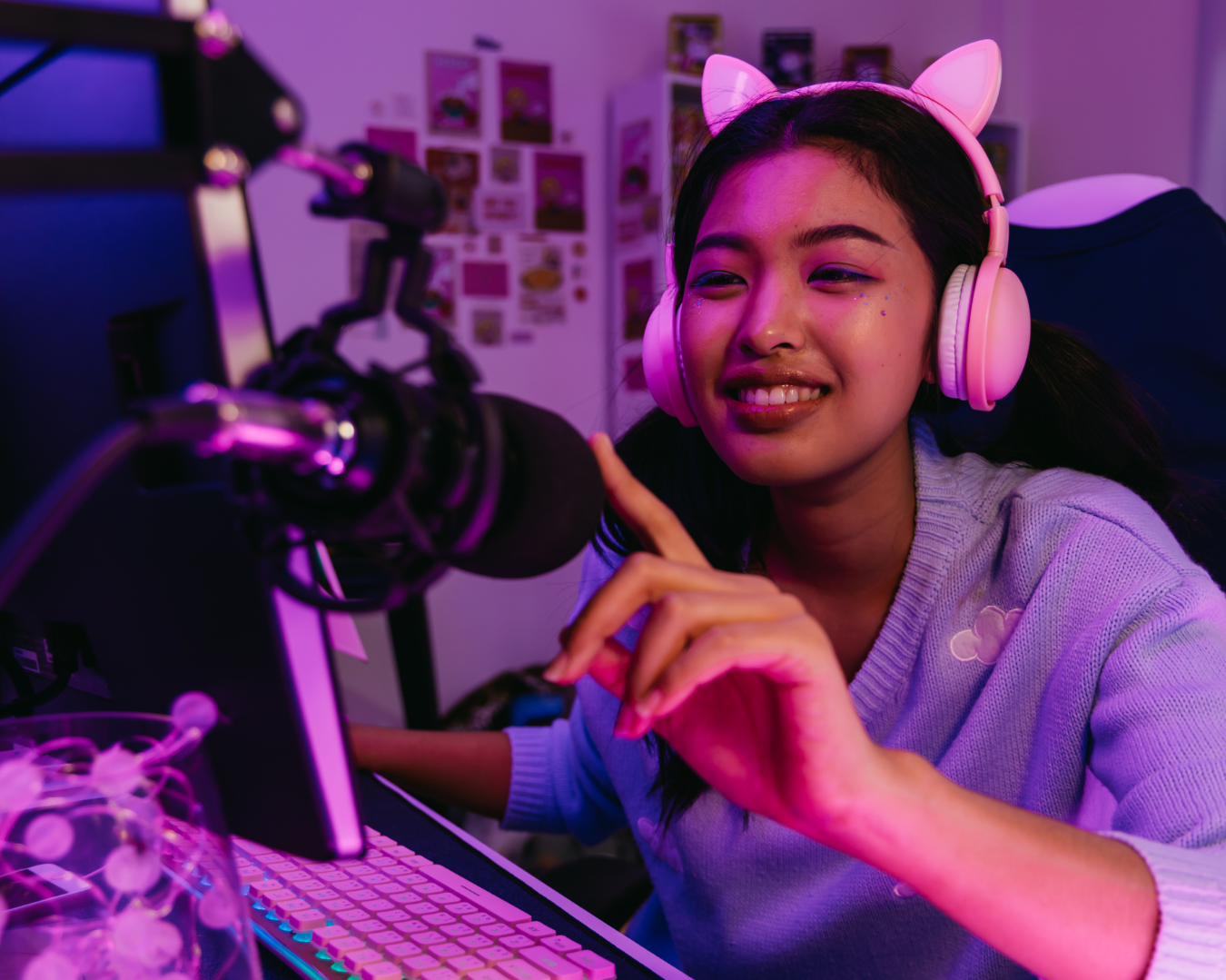 A young woman at her computer, wearing pink cat ear headphones, smiling and pointing at her microphone in a gamer or streamer setup, with colorful lighting and gaming or streaming memorabilia in the background.