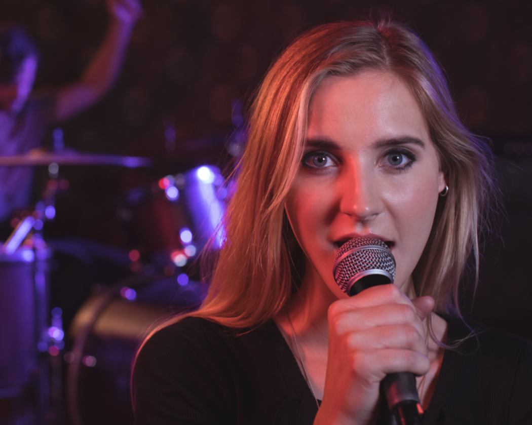 A woman with blonde hair singing into a microphone on stage, with a drum set and blurred band members in the background.
