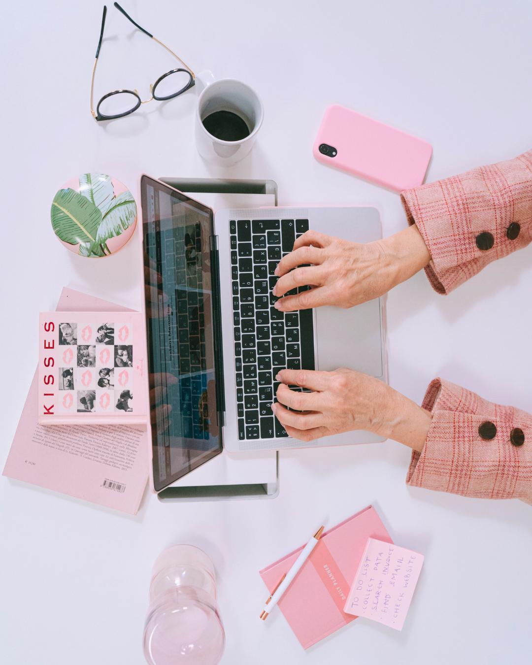 Top-down view of a workspace with a person using a laptop, surrounded by a pink notepad, pen, pink smartphone, mug of coffee, glasses, a pink leaf-patterned container, a pink sticky note, and a glass of water.
