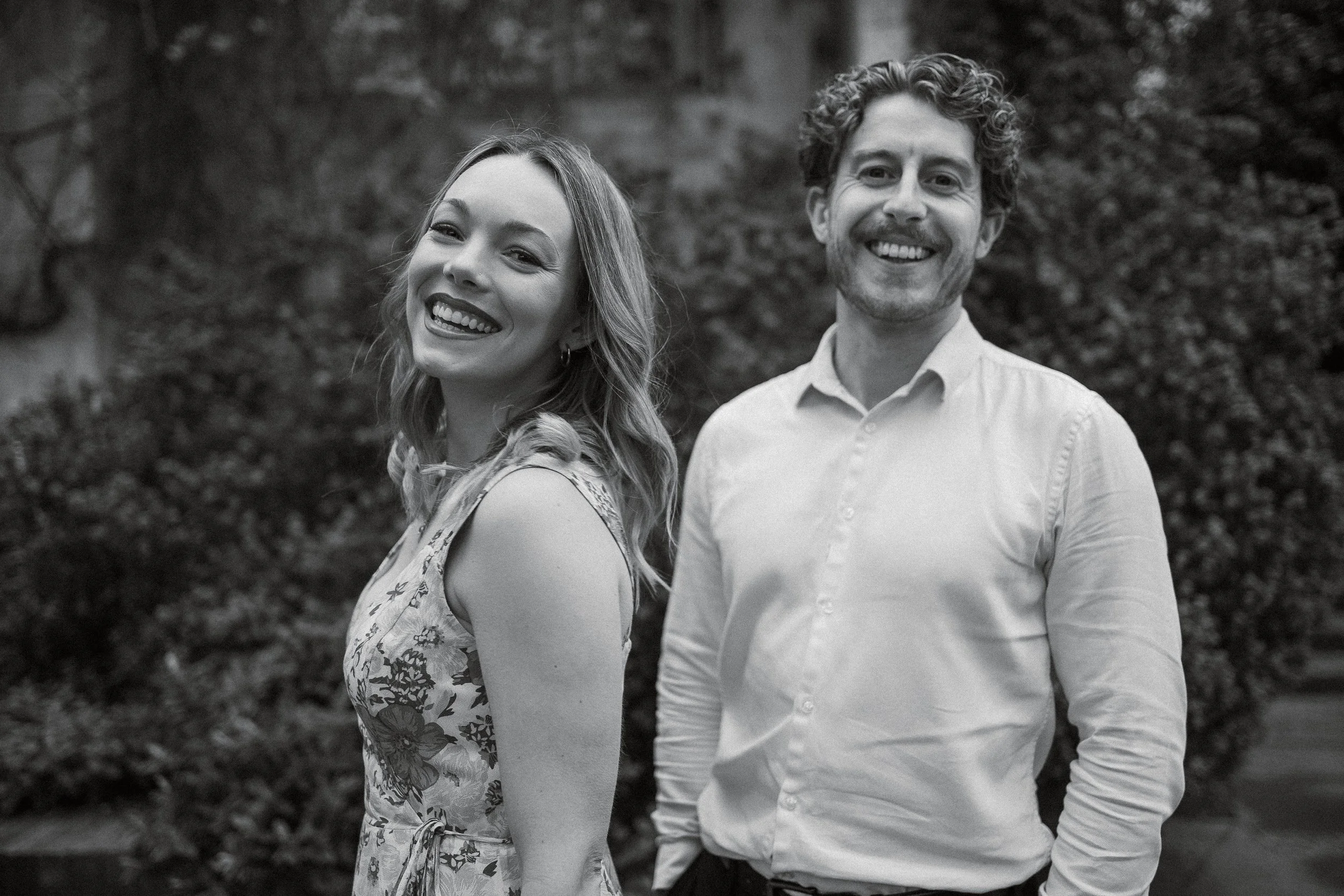 A smiling woman and a smiling man standing outdoors in a garden or park, with greenery in the background.