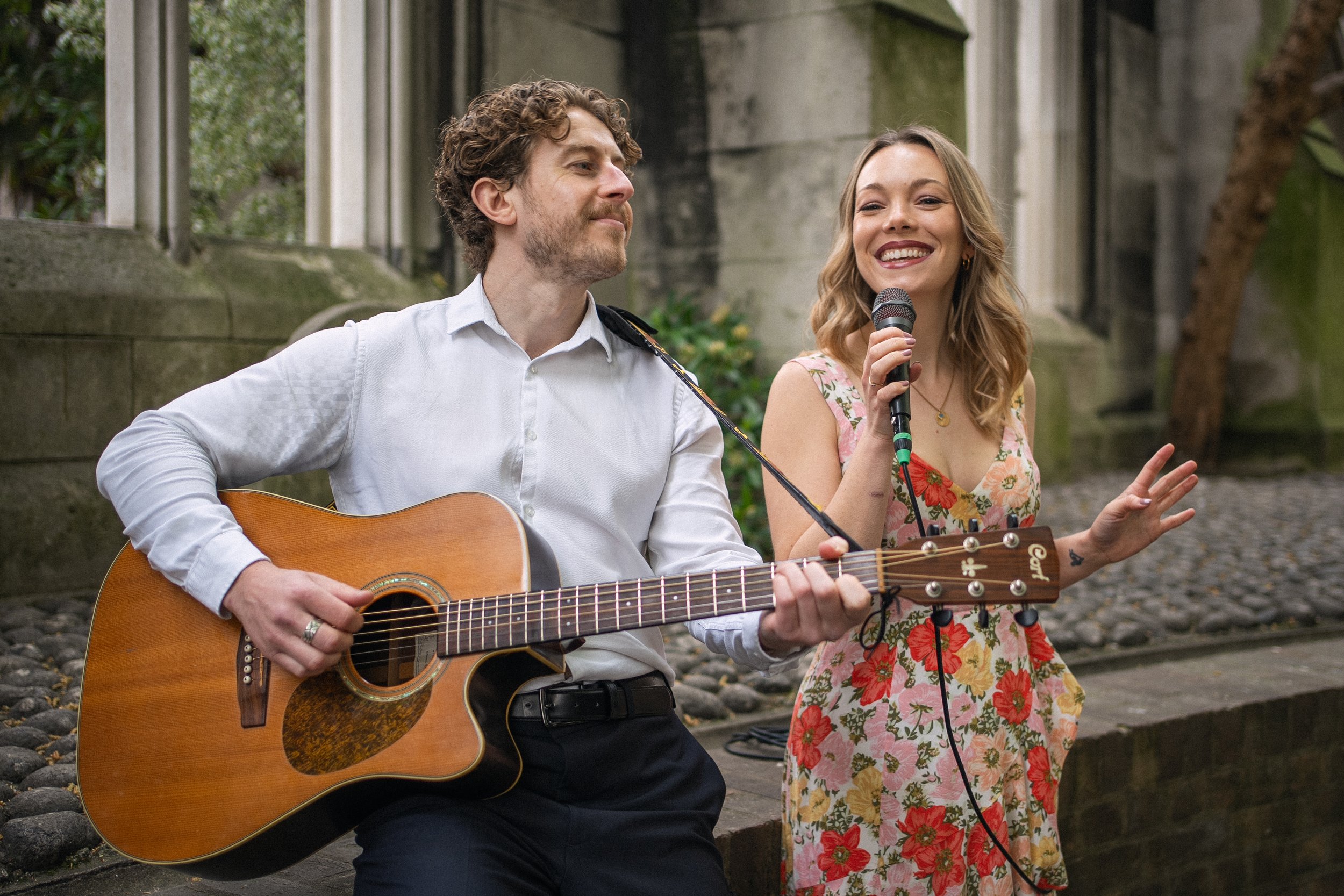 A man playing an acoustic guitar and a woman singing into a microphone outdoors.