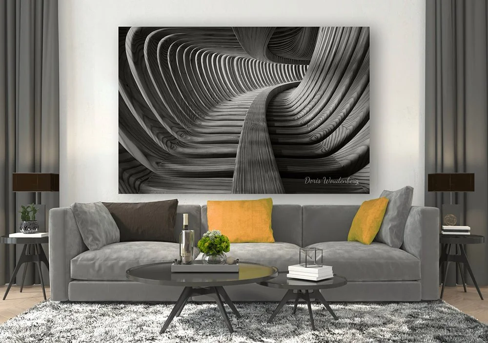 Living room with a large abstract black-and-white artwork of a modern, curving architectural structure hanging above a gray sofa. The sofa has gray, brown, and mustard yellow pillows. There are side tables with lamps on each side, and a round coffee table in front with books, a plant, and decorative items. The room has a textured gray and white rug and light-colored walls.