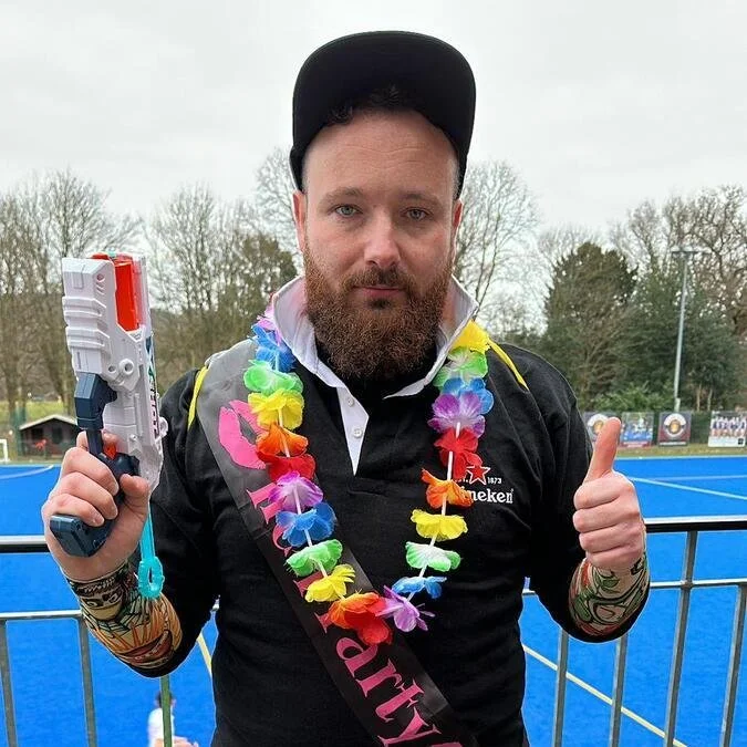 A man with a beard wearing a black cap and a black Heineken shirt, holding a toy gun in one hand and giving a thumbs-up with the other, standing outdoors on a blue sports court with trees in the background.