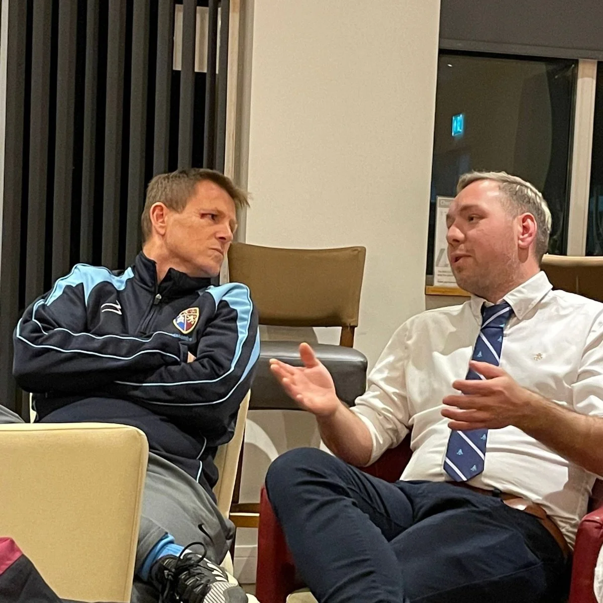 Two men having a serious conversation in a casual indoor setting. One man is wearing a sports jacket, and the other is dressed in a white shirt with a tie.