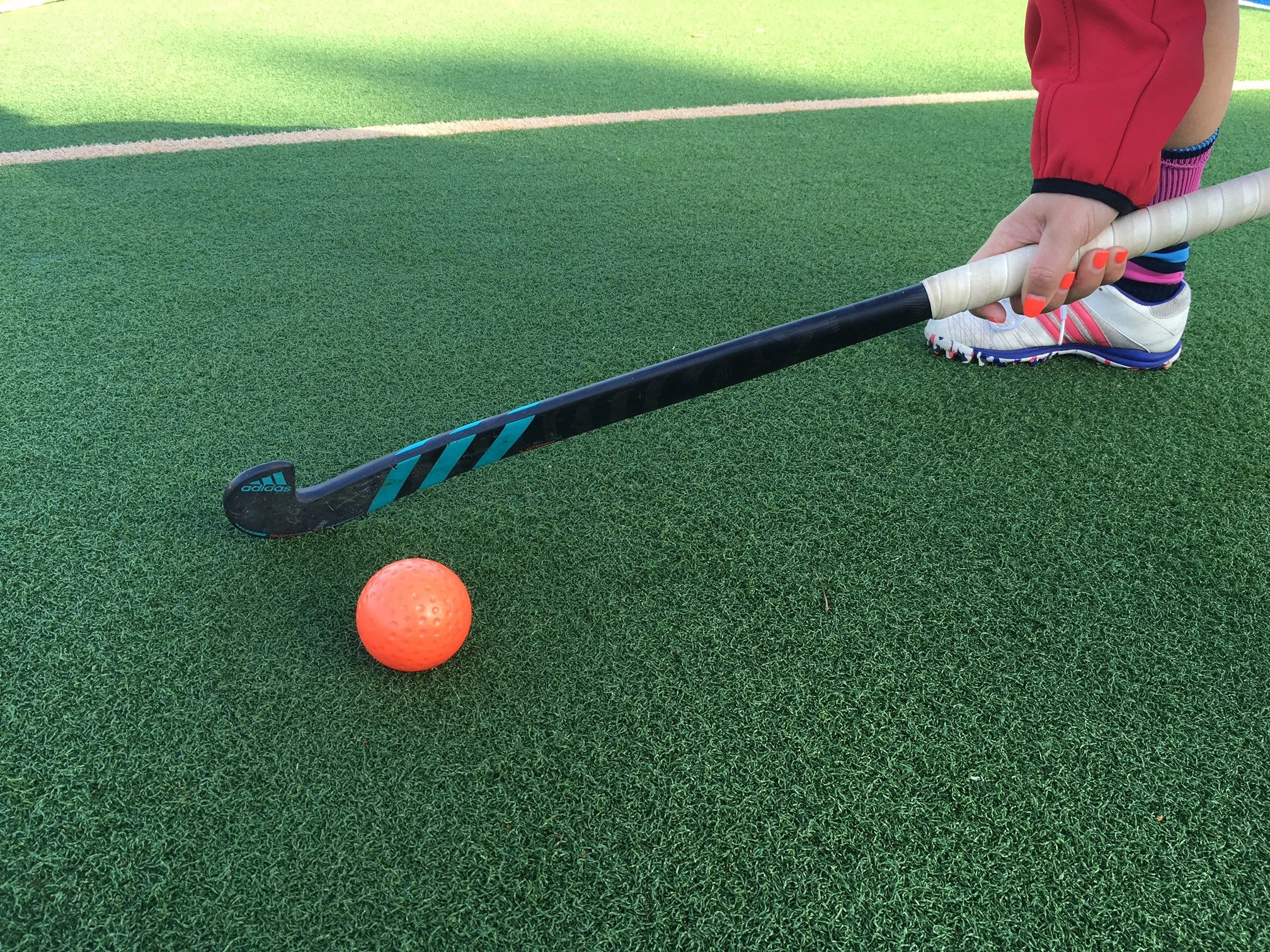 A person with painted nails holding a field hockey stick next to an orange field hockey ball on artificial turf.