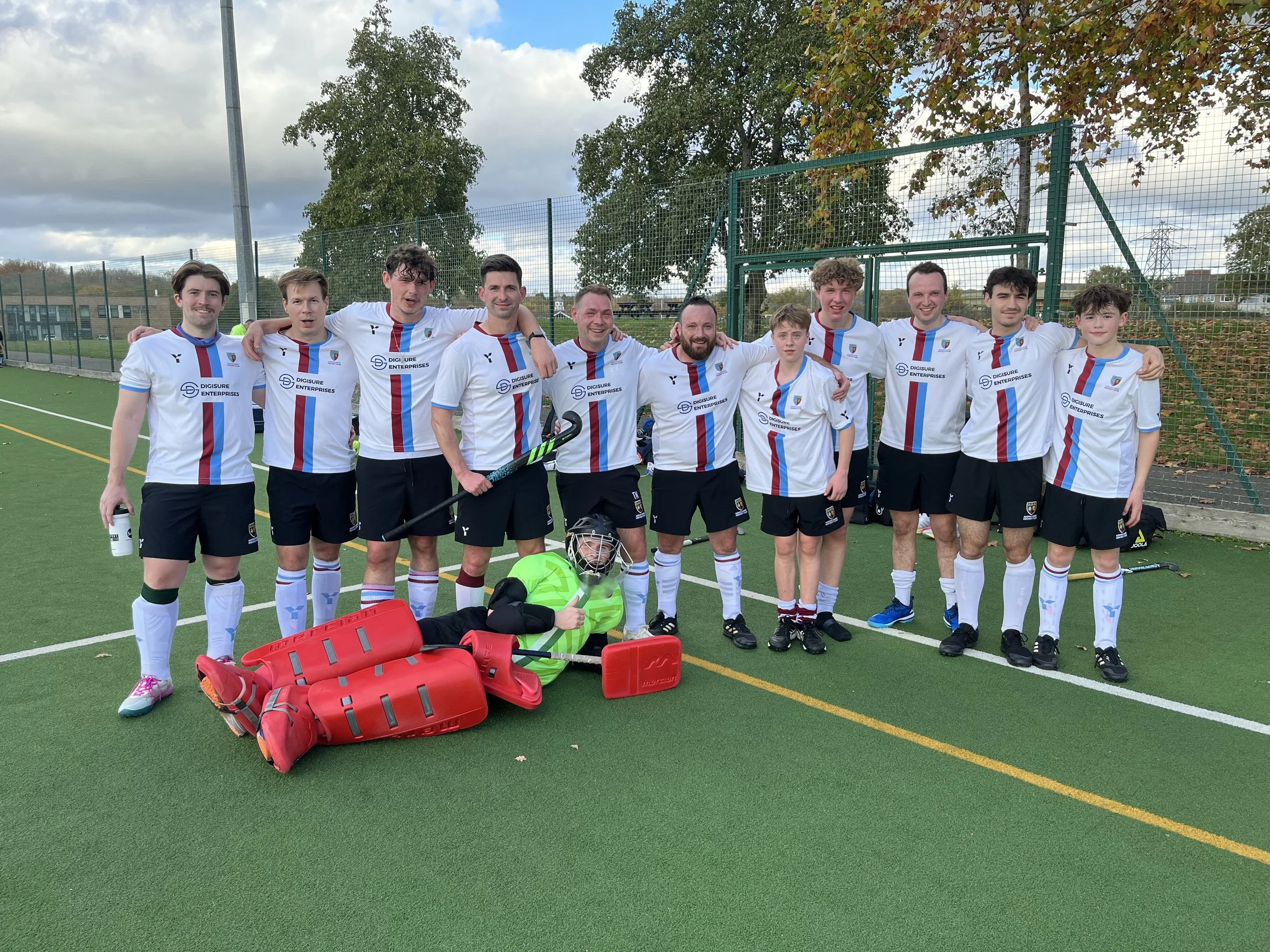A group of hockey players and a young goalie lying on a sports field. They are wearing white jerseys with blue and red stripes and black shorts, smiling for a team photo.