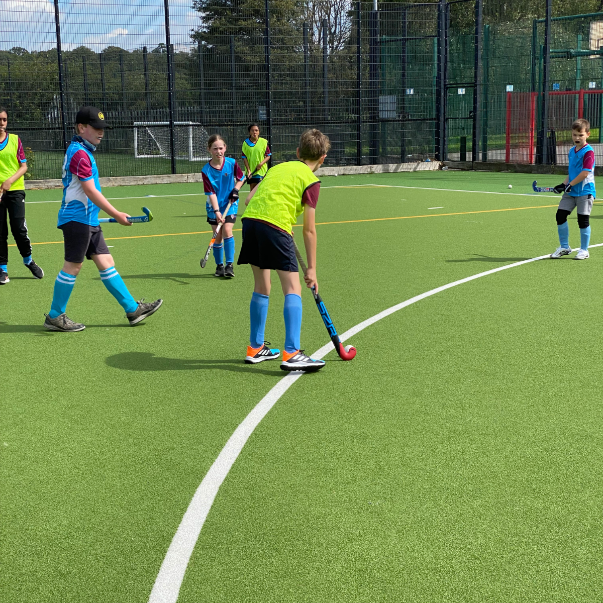 Kids playing field hockey on a green outdoor turf field, with some kids in blue uniforms and others in yellow bibs, all holding hockey sticks and watching a ball.