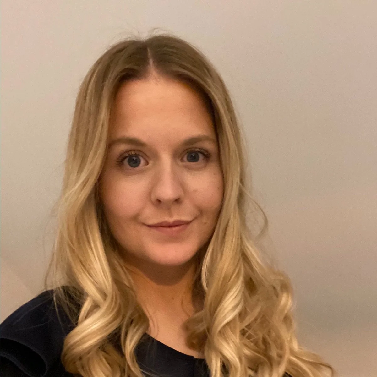 A woman with long blonde hair and blue eyes smiling at the camera against a plain background.
