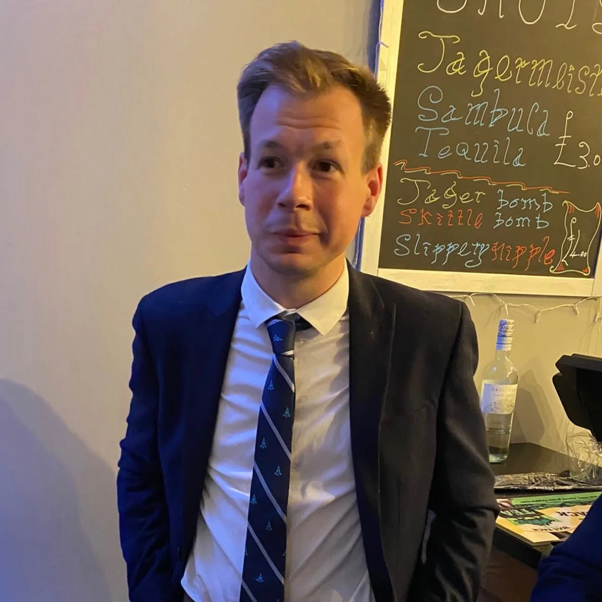 A young man in a suit and tie standing in a restaurant or cafe near a chalkboard menu with food and drink options.