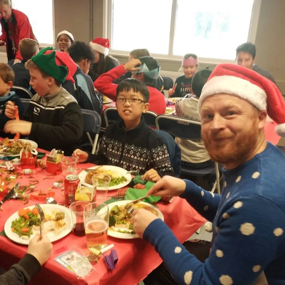 A group of children and an adult wearing Christmas hats gather around a table filled with food at a holiday celebration.