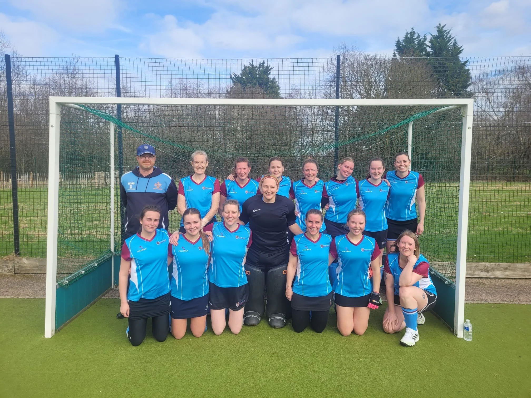 A group of female field hockey players and a coach posing for a team photo in front of a goal on the field, with a water bottle on the ground nearby.