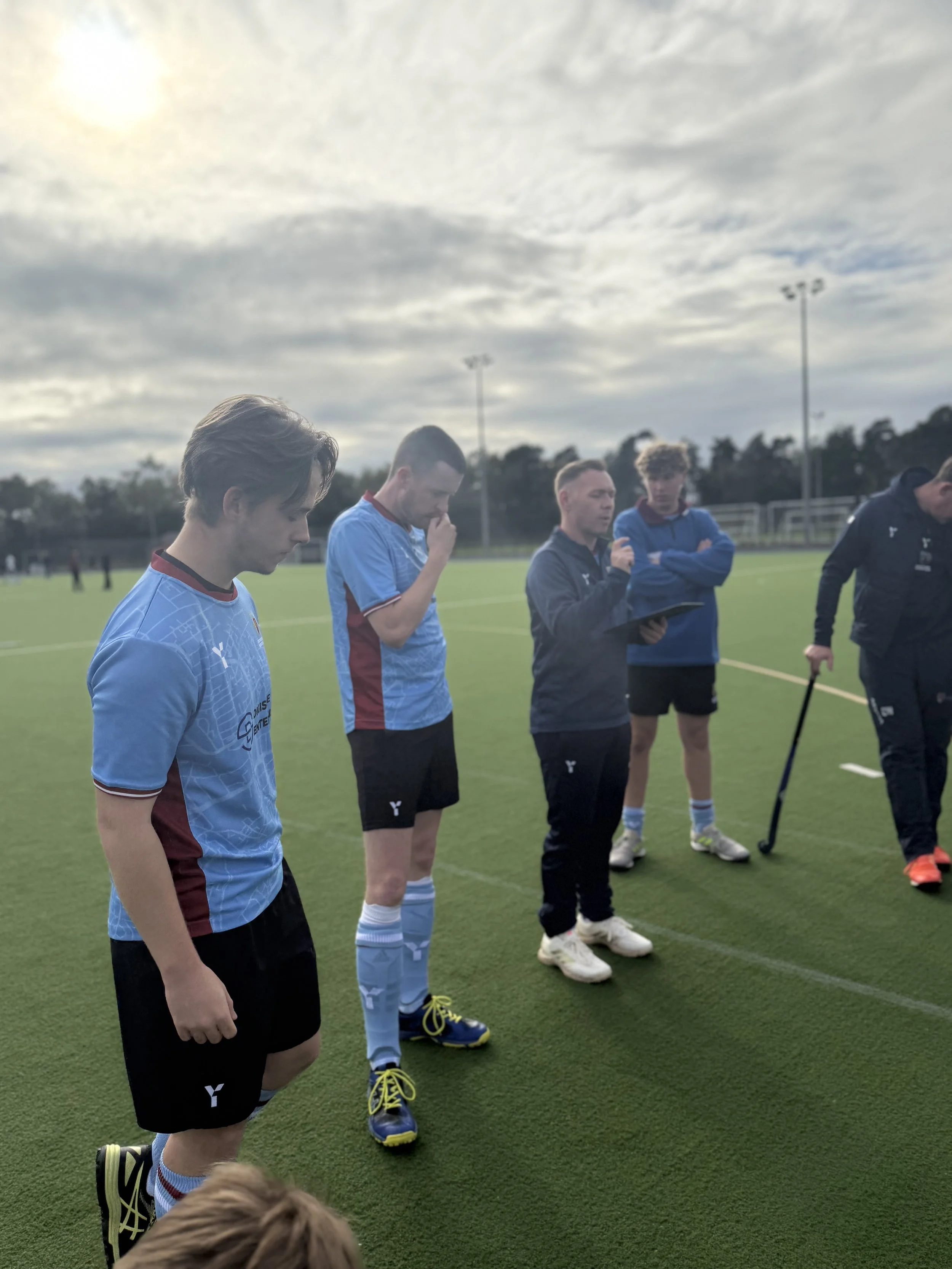 A group of young men in sports uniforms standing on a soccer field, with a coach and officials, under a cloudy sky.