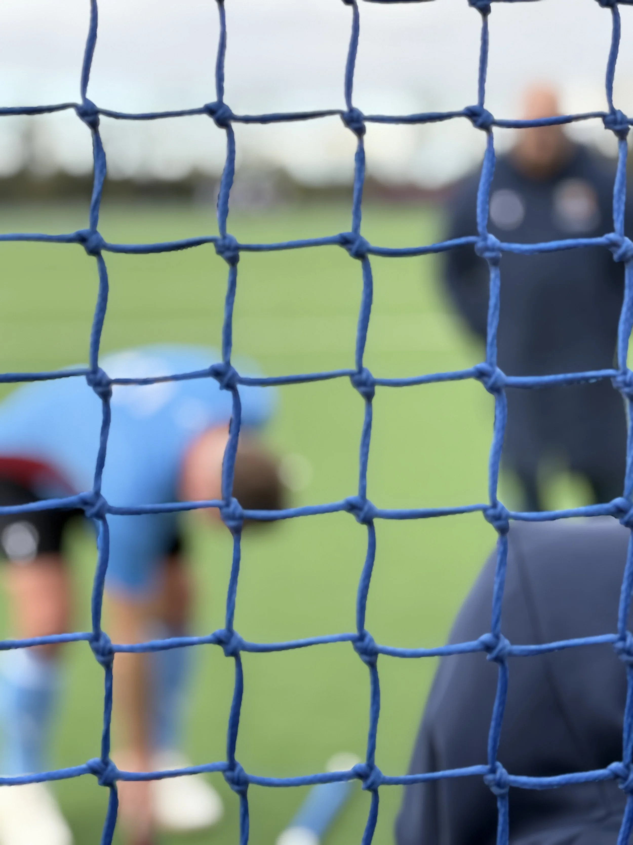 Close-up of a blue sports net on a field, with two people and a child playing in the background, slightly out of focus.