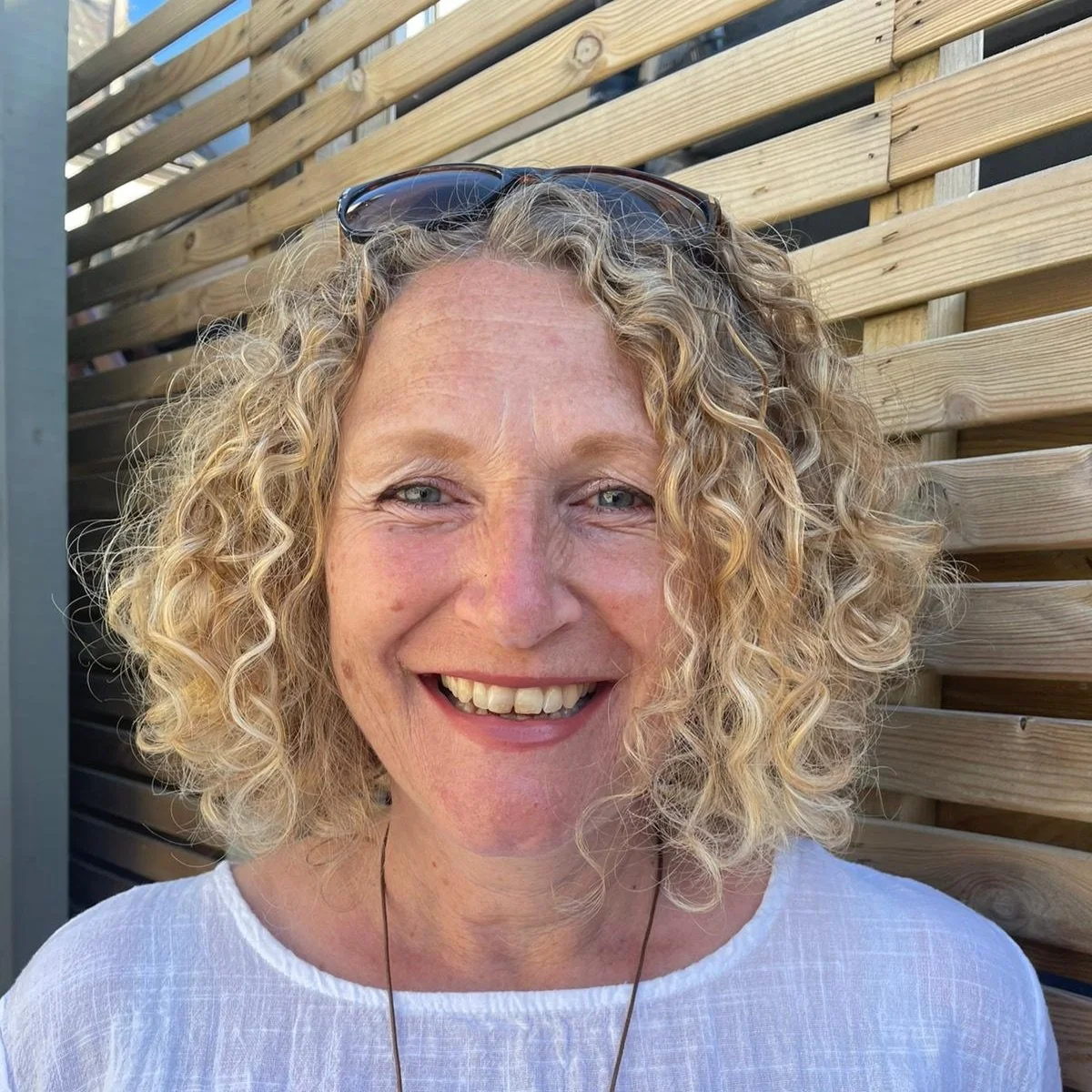 Close-up of a smiling woman with curly blonde hair, wearing sunglasses on her head, in front of a wooden fence.
