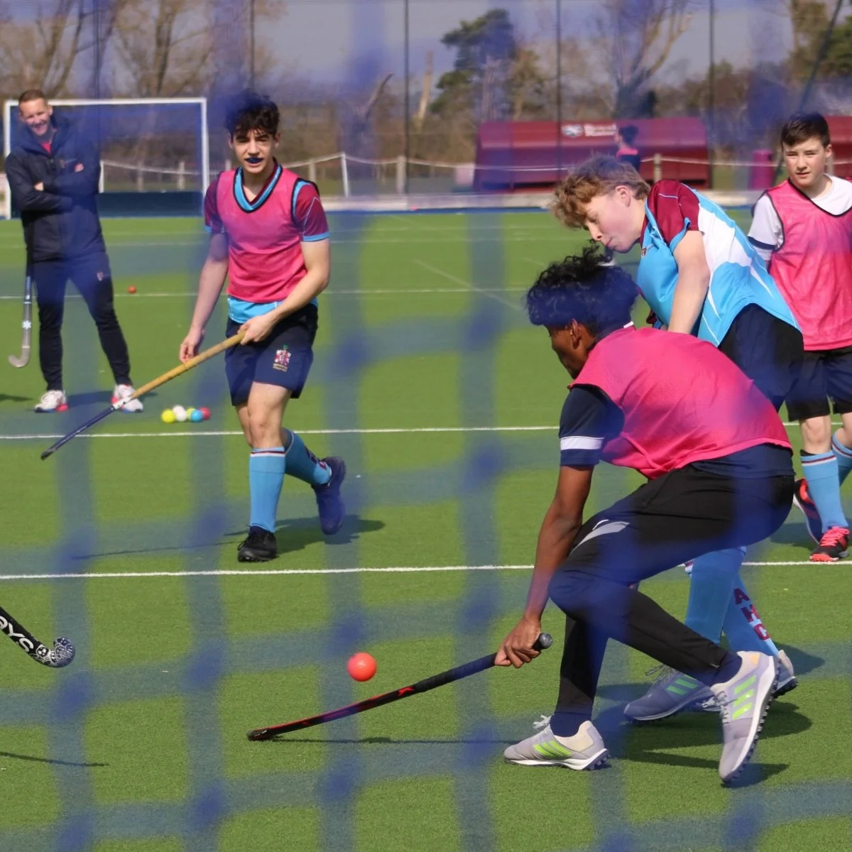Young male field hockey players practicing on a green turf field, with coach and other players observing in the background.