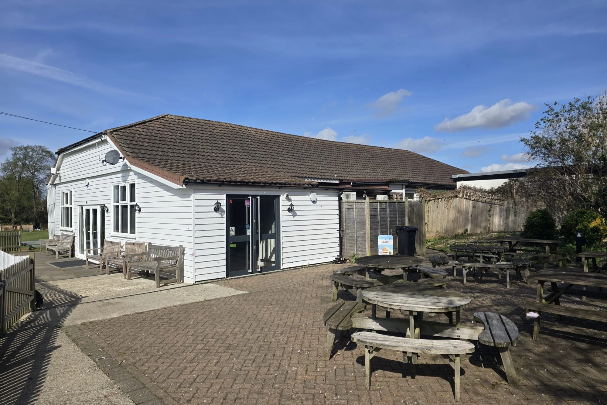 A white building with a brown sloped roof, outdoor benches along the wall, and wooden picnic tables on a brick and concrete patio, under a partly cloudy sky.