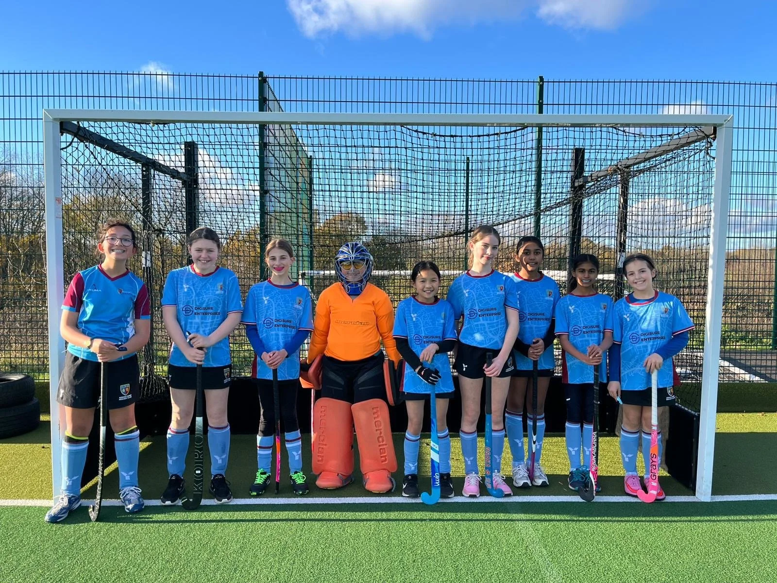 Group of nine young field hockey players, including a goalkeeper in orange, standing in front of a goal on a green turf field under a blue sky.