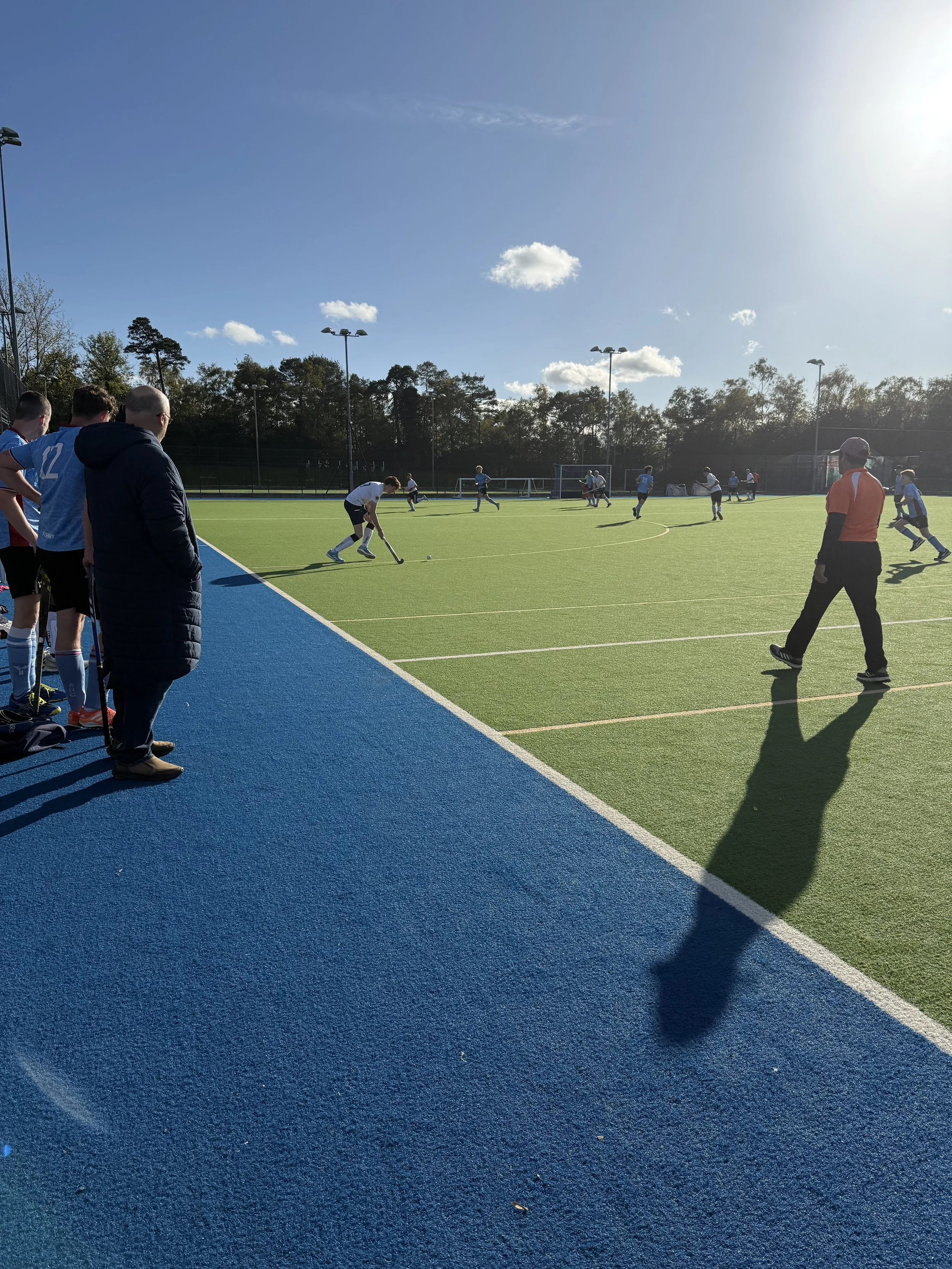 A field hockey game with players on a green turf field, a coach or referee in a red shirt walking on the sideline, and a group of players watching near the blue track under a clear sky with some clouds.