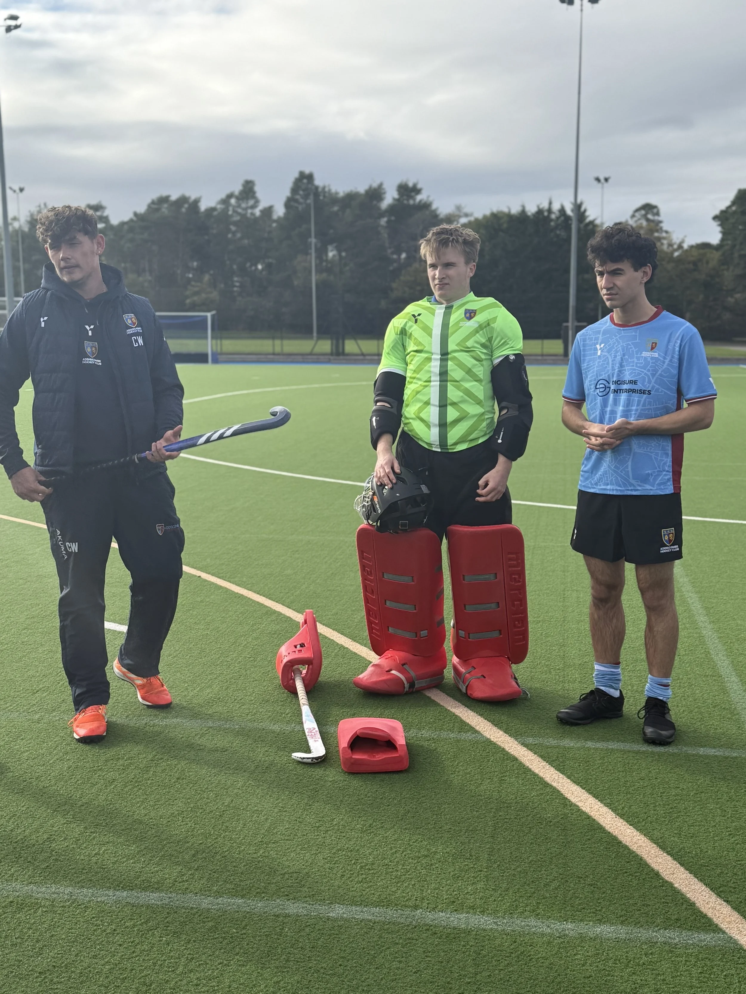 Three individuals standing on a sports field, with one dressed as a field hockey goalkeeper wearing protective gear, and the others in sportswear, holding field hockey equipment.
