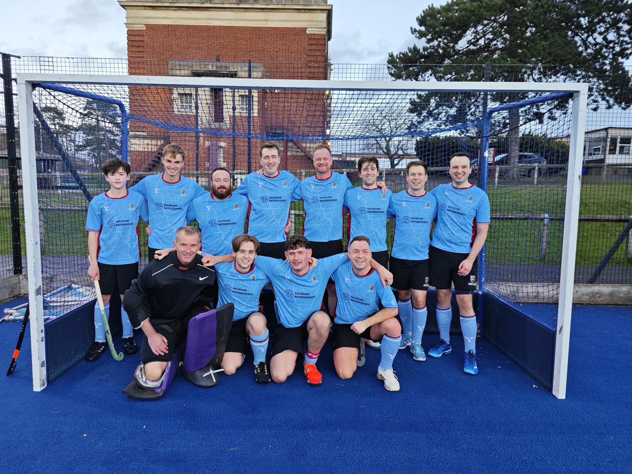 A group of eleven male field hockey players in matching light blue jerseys and black shorts, standing inside a goal on a blue sports field, posing for a team photo with some players kneeling in front and others standing behind.