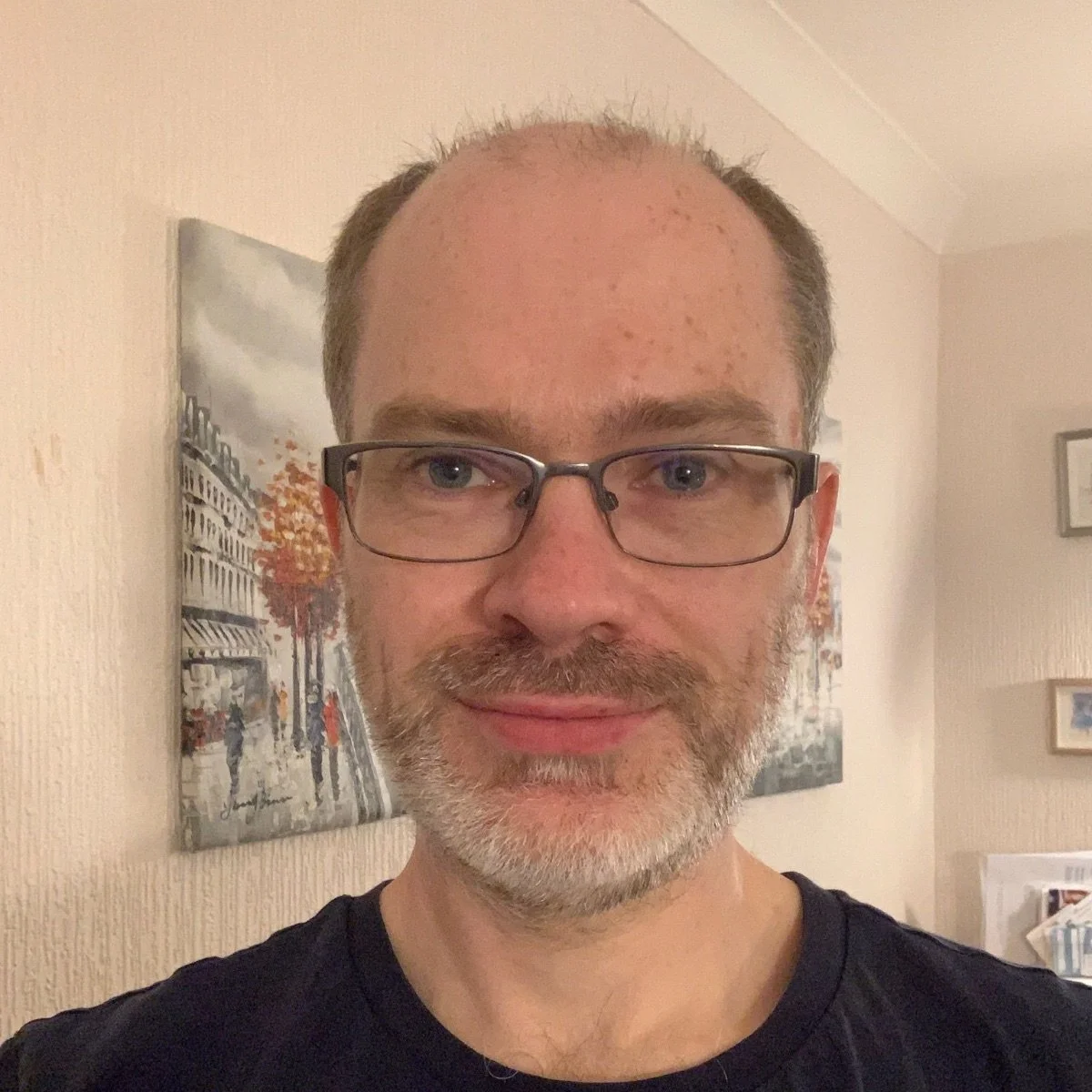 A man with glasses and a beard smiling at the camera indoors with artwork on the wall behind him.