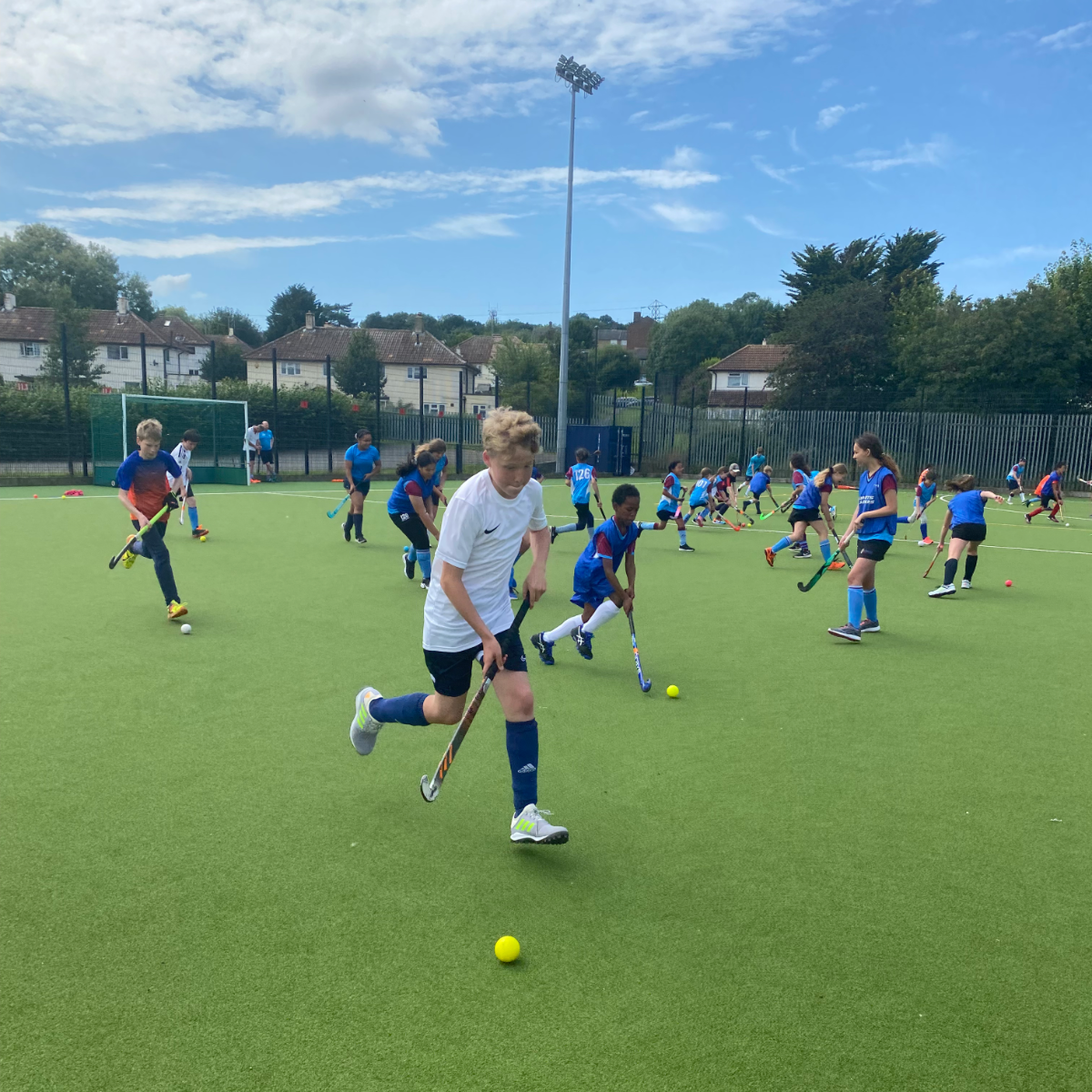 Children playing field hockey on an outdoor turf field under a partly cloudy sky.