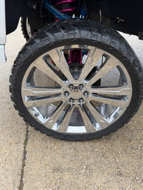 Close-up of a car wheel with a large chrome rim and a rugged tire. The pink suspension components are visible behind the wheel.