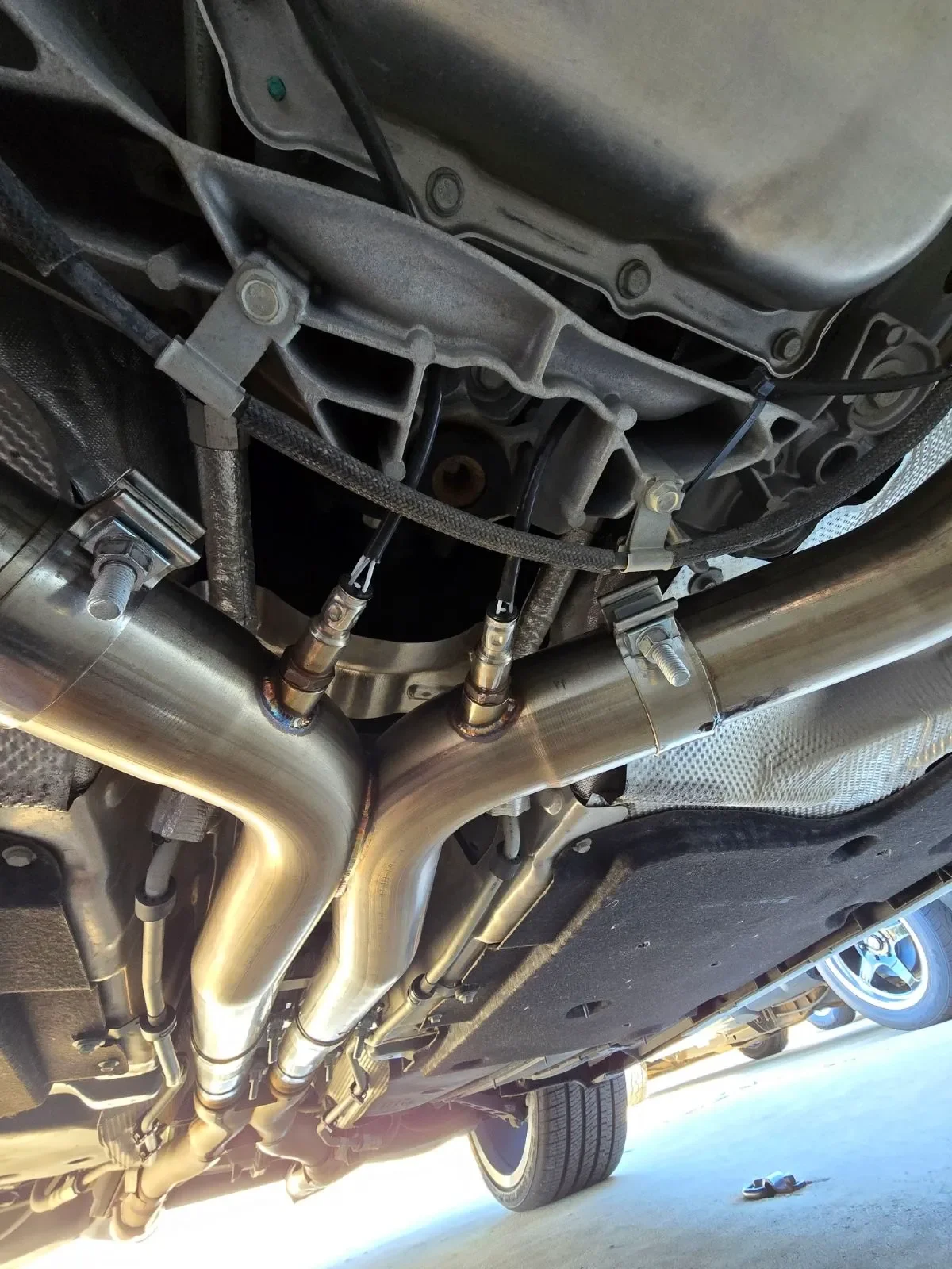 Underneath view of a car showing metallic exhaust pipes connected to the engine with heat shields and mounted to the underside of the vehicle, with part of a tire visible in the background.