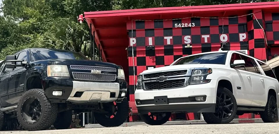Two Chevrolet vehicles, a black truck with off-road tires and a white sedan with black wheels, parked in front of a red and black checkered structure with a sign reading 'PIT STOP'.