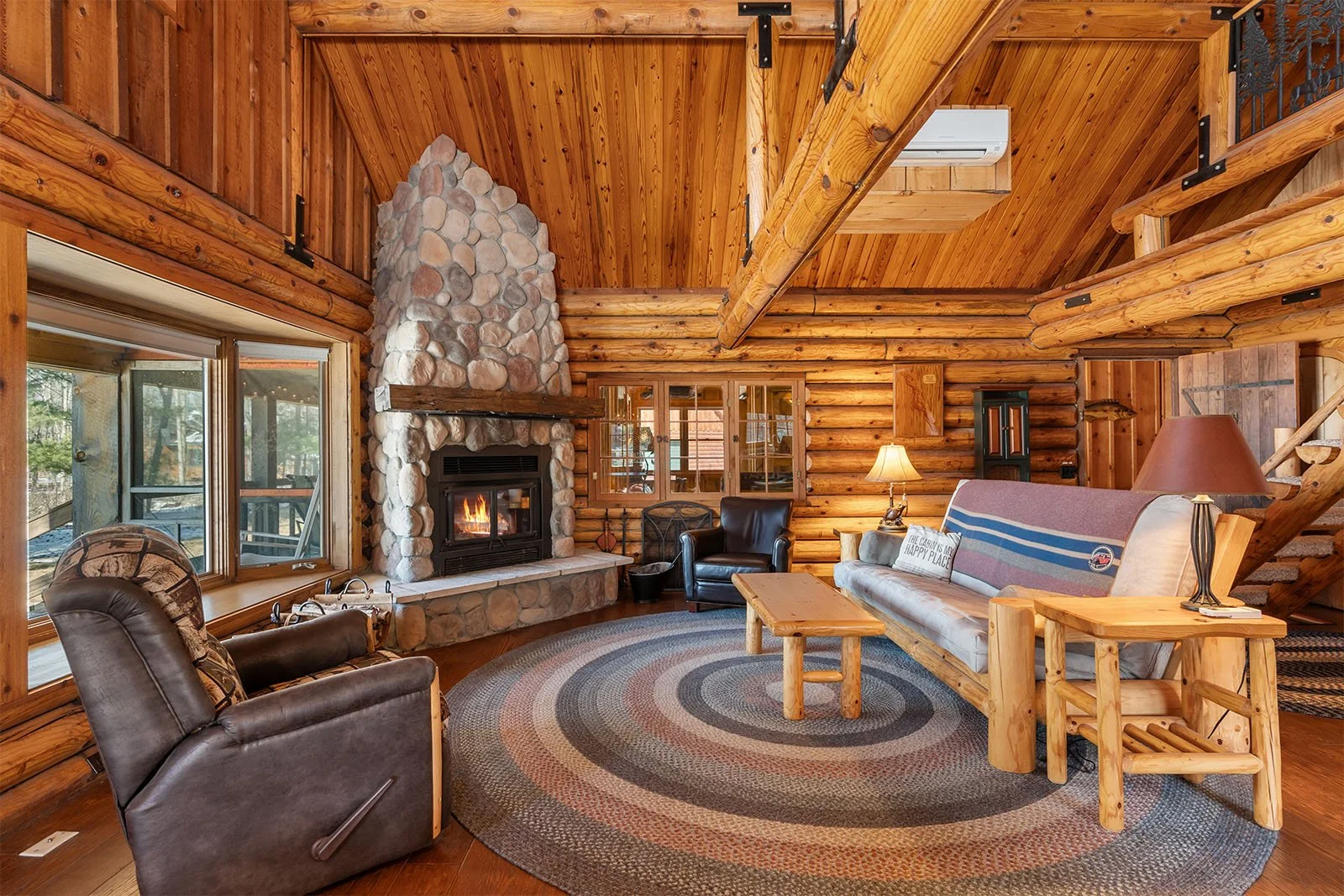 Cozy log cabin living room with a stone fireplace, wooden walls, and furniture including a leather armchair, a sofa with a blanket, a wooden coffee table, rugs, and lamps.