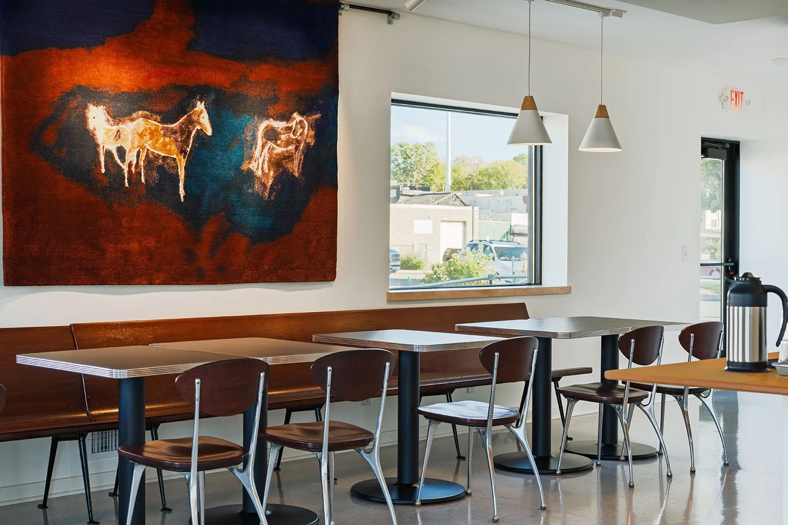 Interior of a modern cafe with a row of wooden chairs and tables, a large painting of horses on the wall, a window showing parked cars outside, and pendant lights hanging from the ceiling.