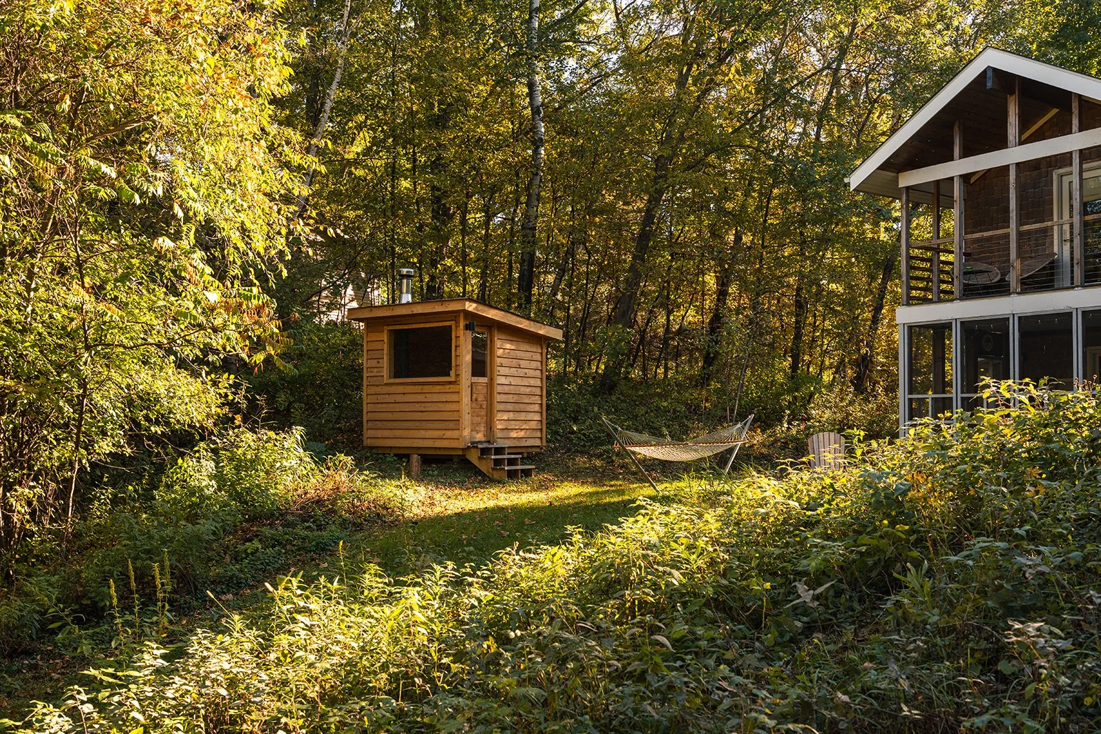 A backyard scene during daytime showing a sauna, a hammock, a cabin with a screened porch, surrounded by trees with fall foliage.