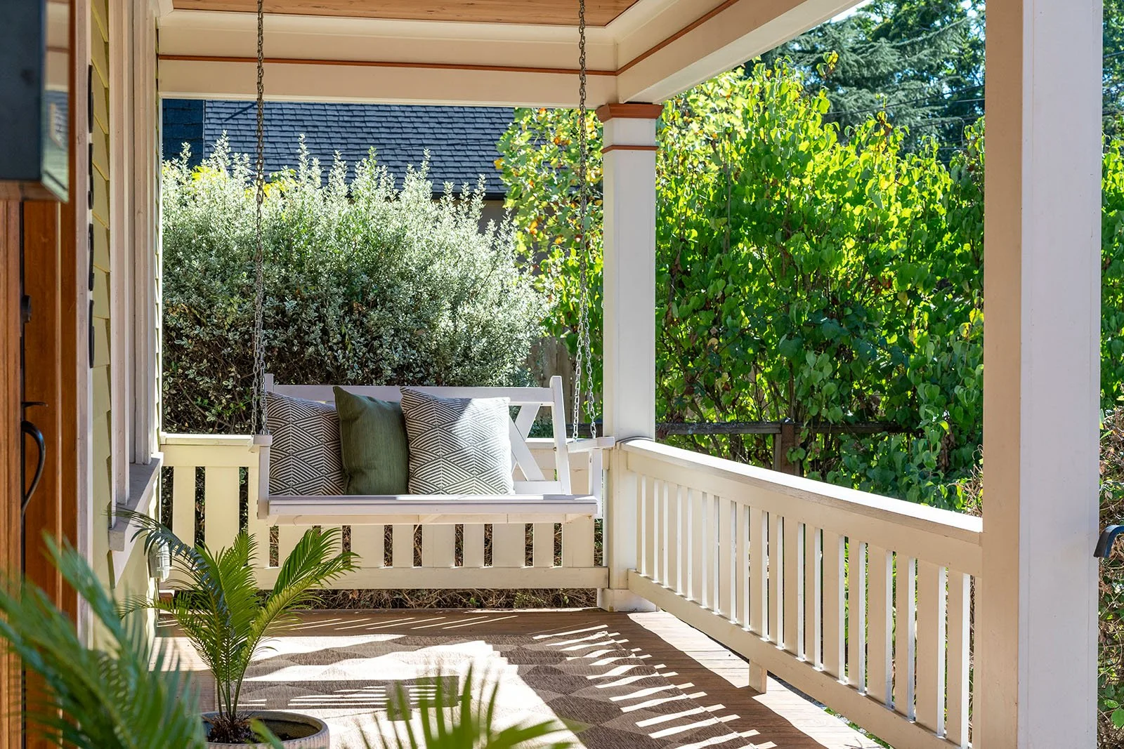 A cozy porch with a white porch swing with three decorative pillows, surrounded by greenery and plants, with sunlight casting shadows on the wooden floor.