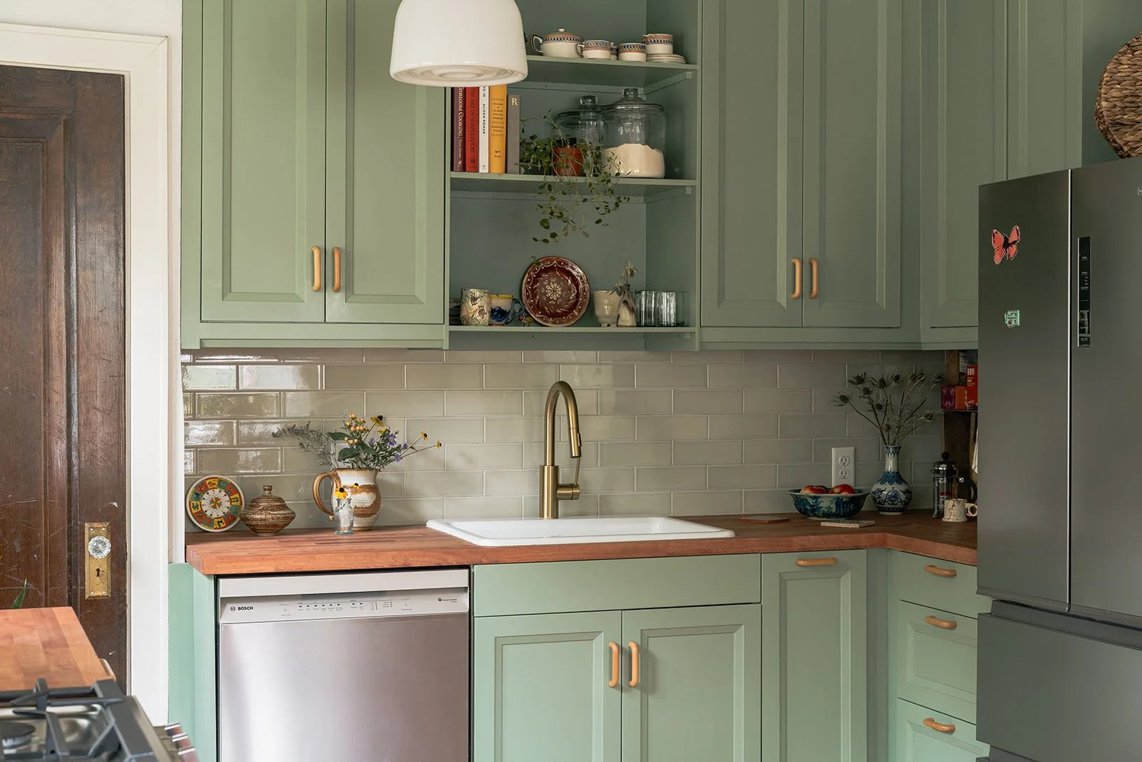 Kitchen with light green cabinets, a white farmhouse sink with a brass faucet, and a brick backsplash. There are open shelves with dishes, glasses, and jars, along with decorative items and plants. The countertop is wood.