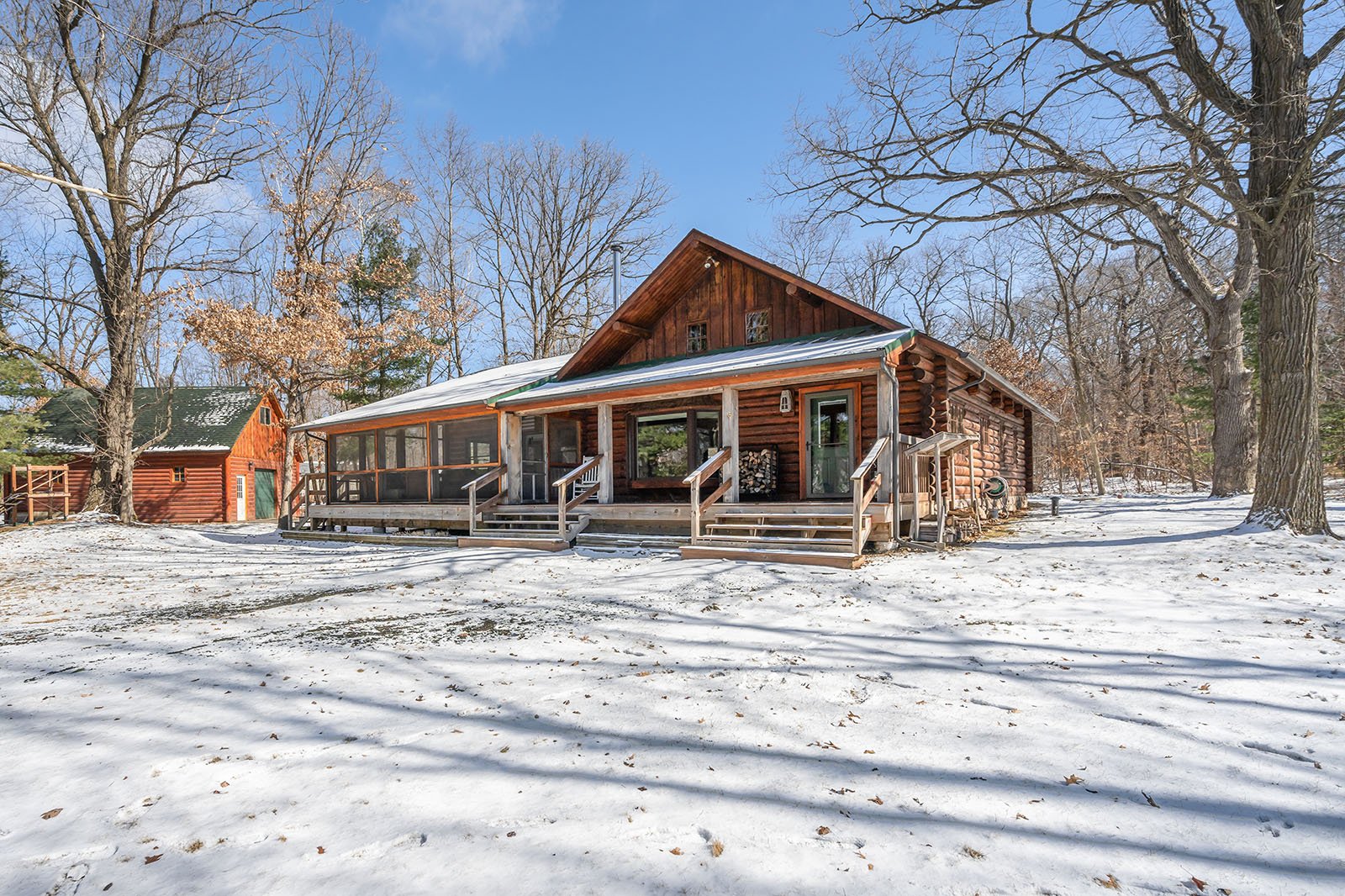 A wooden cabin with a screened porch and garage nearby, surrounded by snow-covered ground and leafless trees under a clear blue sky.
