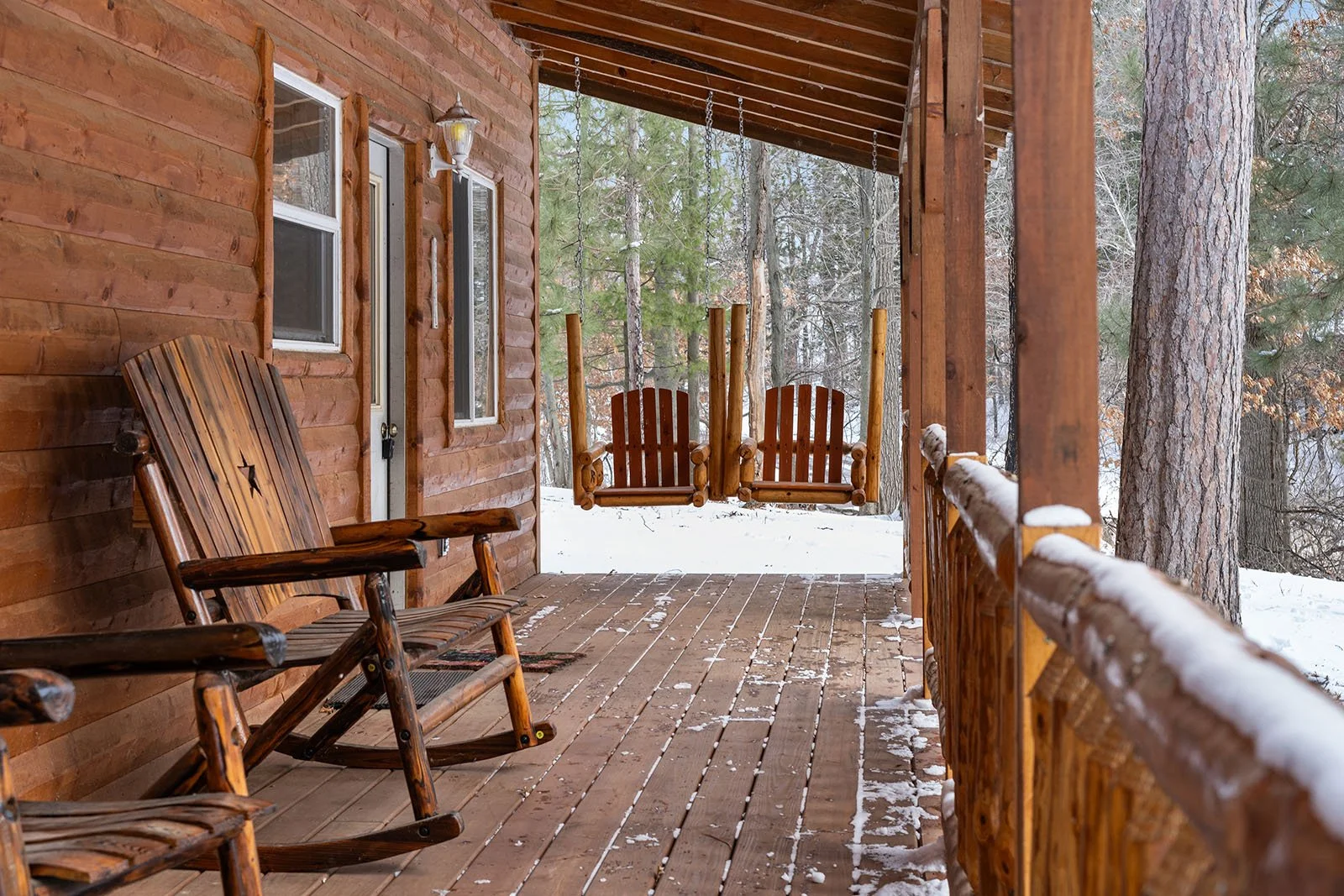 Wooden porch of a log cabin with a rocking chair and two swaying wooden swings, snow on the ground and forest trees in the background.