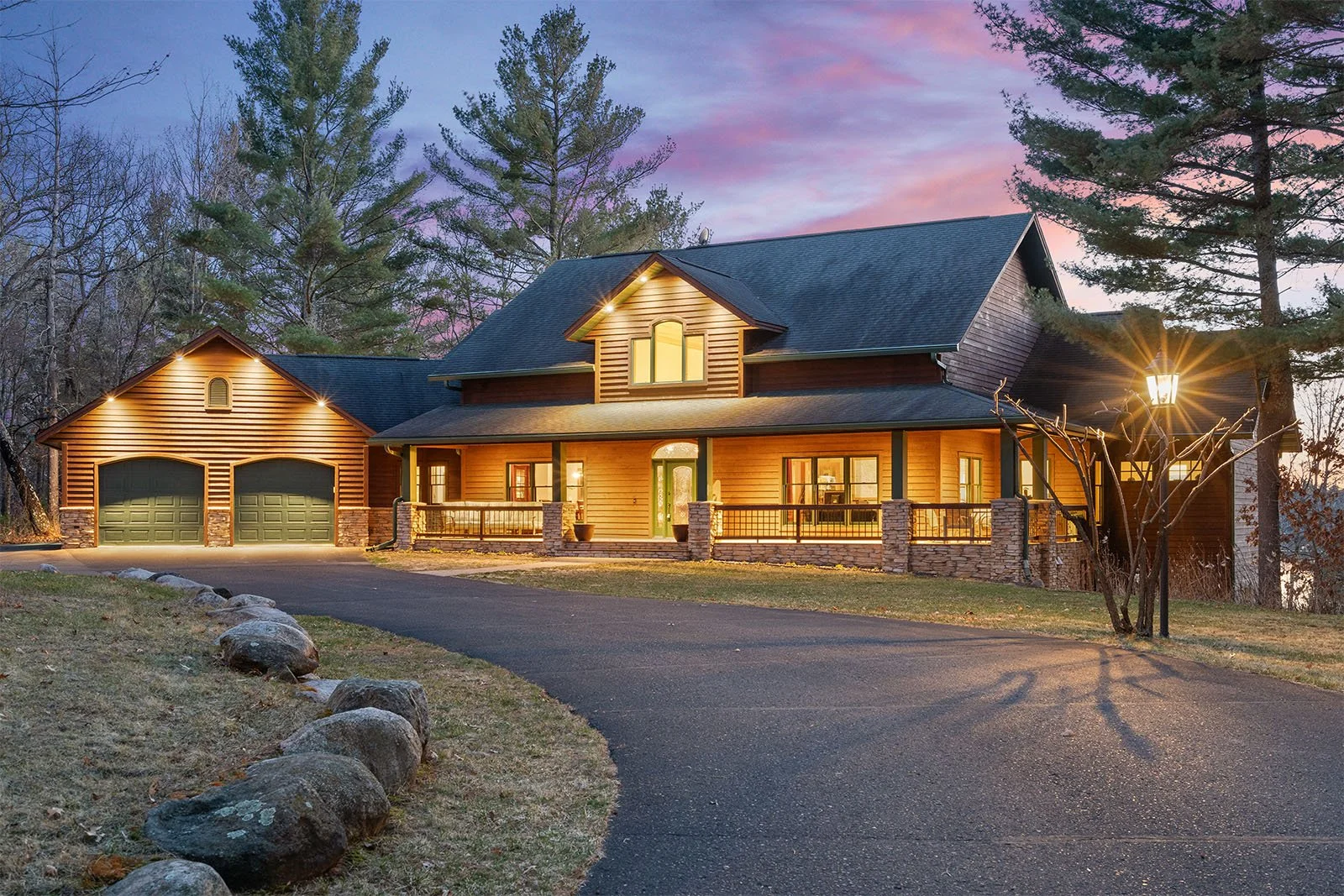 A large house with wood siding, a front porch, and a garage, illuminated at dusk with outdoor lighting, surrounded by trees and a stone-lined driveway.
