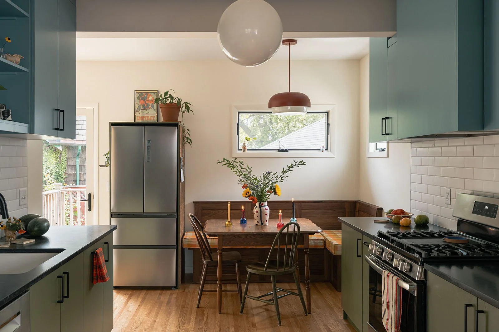 Kitchen with green cabinets, stainless steel refrigerator, white subway tile backsplash, black countertops, wooden floor, small dining nook with a wooden table, floral arrangement, and four candles, two hanging pendant lights, and a door and window l