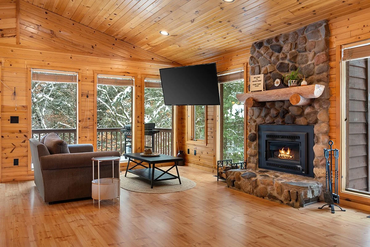Living room with wood-paneled walls and ceiling, large windows showing snowy trees outside, a stone fireplace with a wooden mantel, a mounted flat-screen TV, a gray sofa, a round side table, and a black coffee table on a round rug.