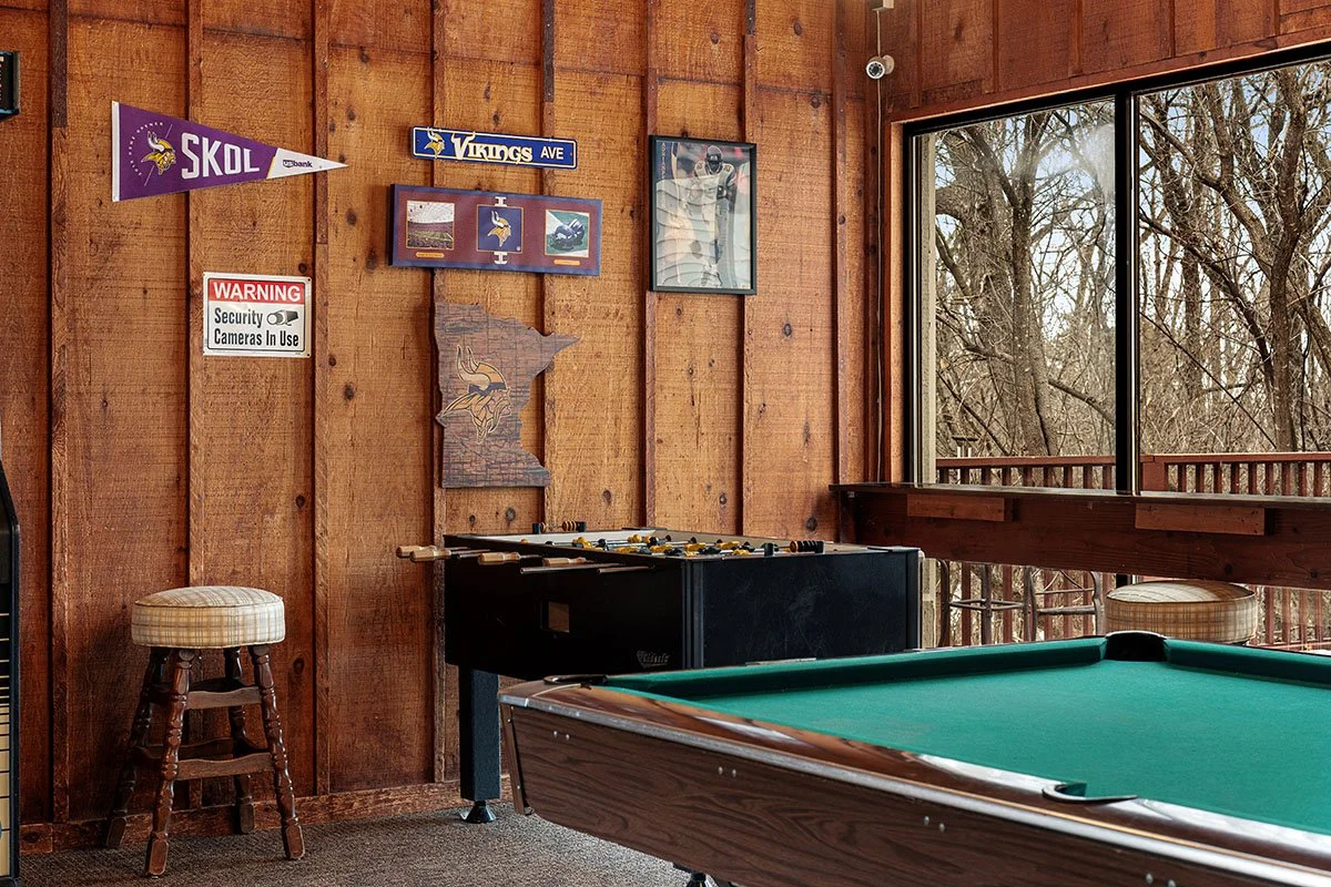 A game room with a pool table, foosball table, wooden walls decorated with Minnesota Vikings memorabilia, and a large window showing trees outside.