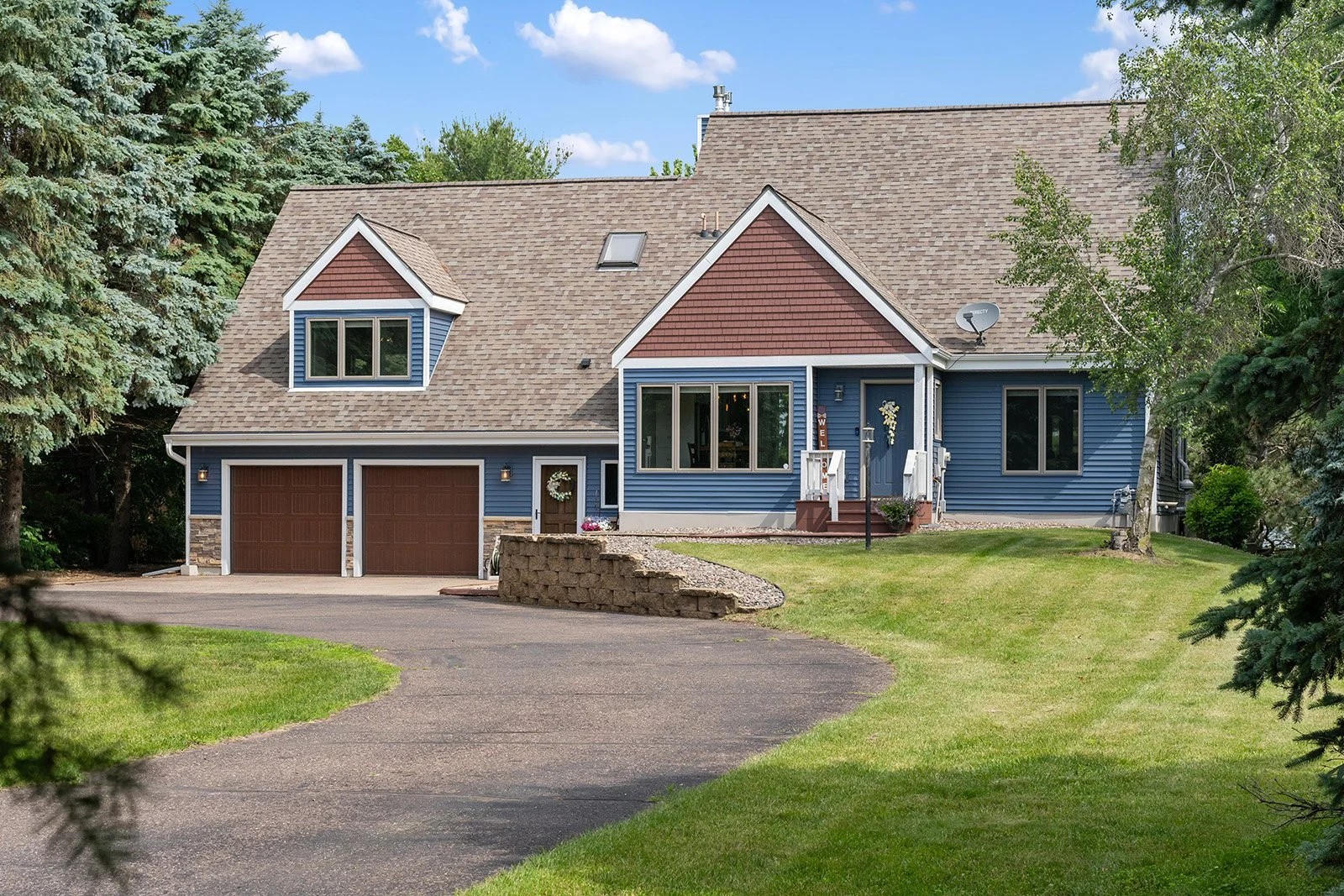 A suburban house with a blue exterior, brown garage doors, and a well-maintained lawn, surrounded by trees and a paved driveway.