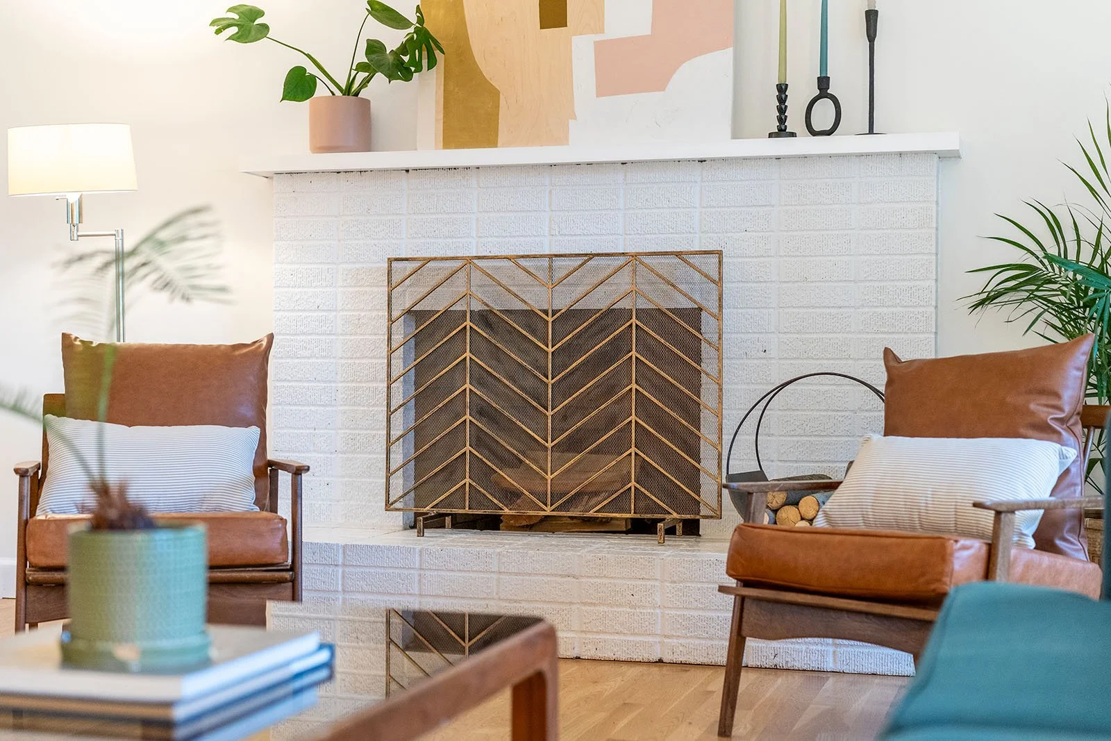 Living room with two brown leather armchairs, a white brick fireplace with a decorative metal screen, a white mantle with plants and art, and a wooden coffee table with a plant and books, potted plants and candles.